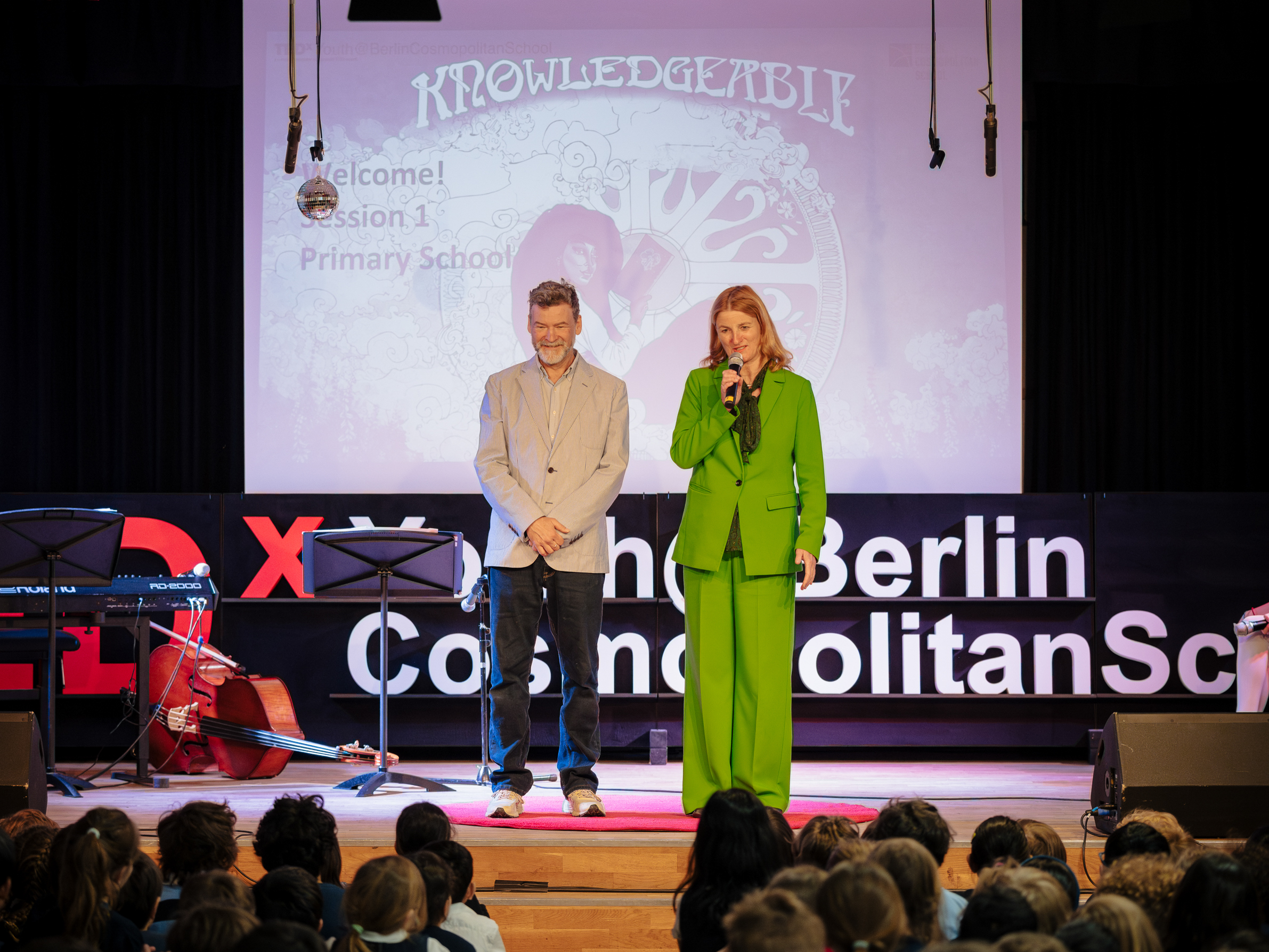 Cosmopolitanschool Berlin Ein Mann und eine Frau stehen auf der Bühne und sprechen bei einer TEDxYouth-Veranstaltung zum Publikum. Auf der Leinwand hinter ihnen erscheint das Wort KNOWLEDGERBLZ und "Welcome! Session 1 Primary School". Auf der linken Seite sind Musikinstrumente zu sehen. loading=