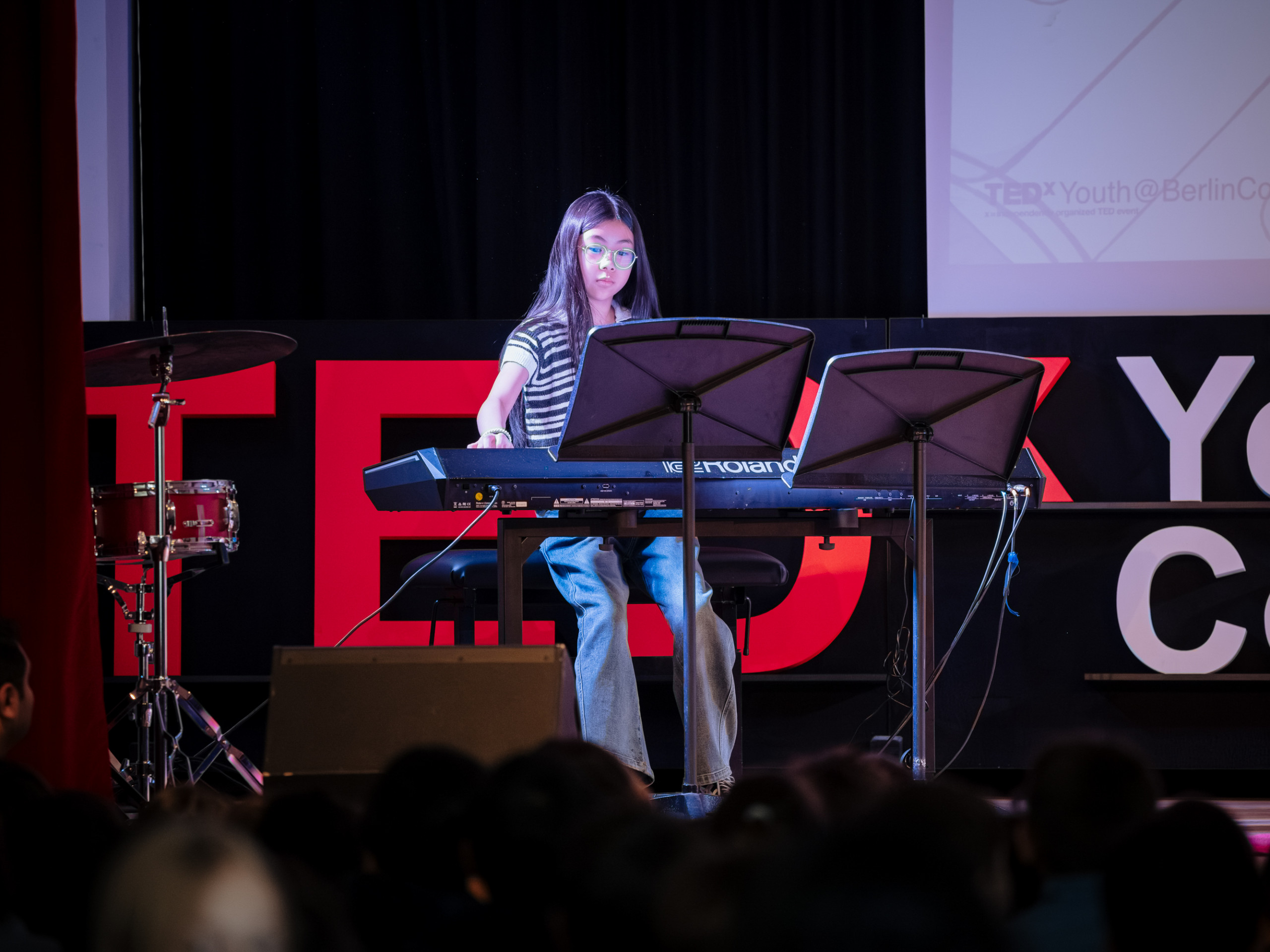 Cosmopolitanschool Berlin Eine junge Frau spielt auf der Bühne einer TEDxYouth-Veranstaltung ein Keyboard und liest Noten. Neben ihr steht ein Schlagzeug, und im Hintergrund ist ein großes TEDx-Schild zu sehen. Im Vordergrund schaut ein Publikum zu. loading=