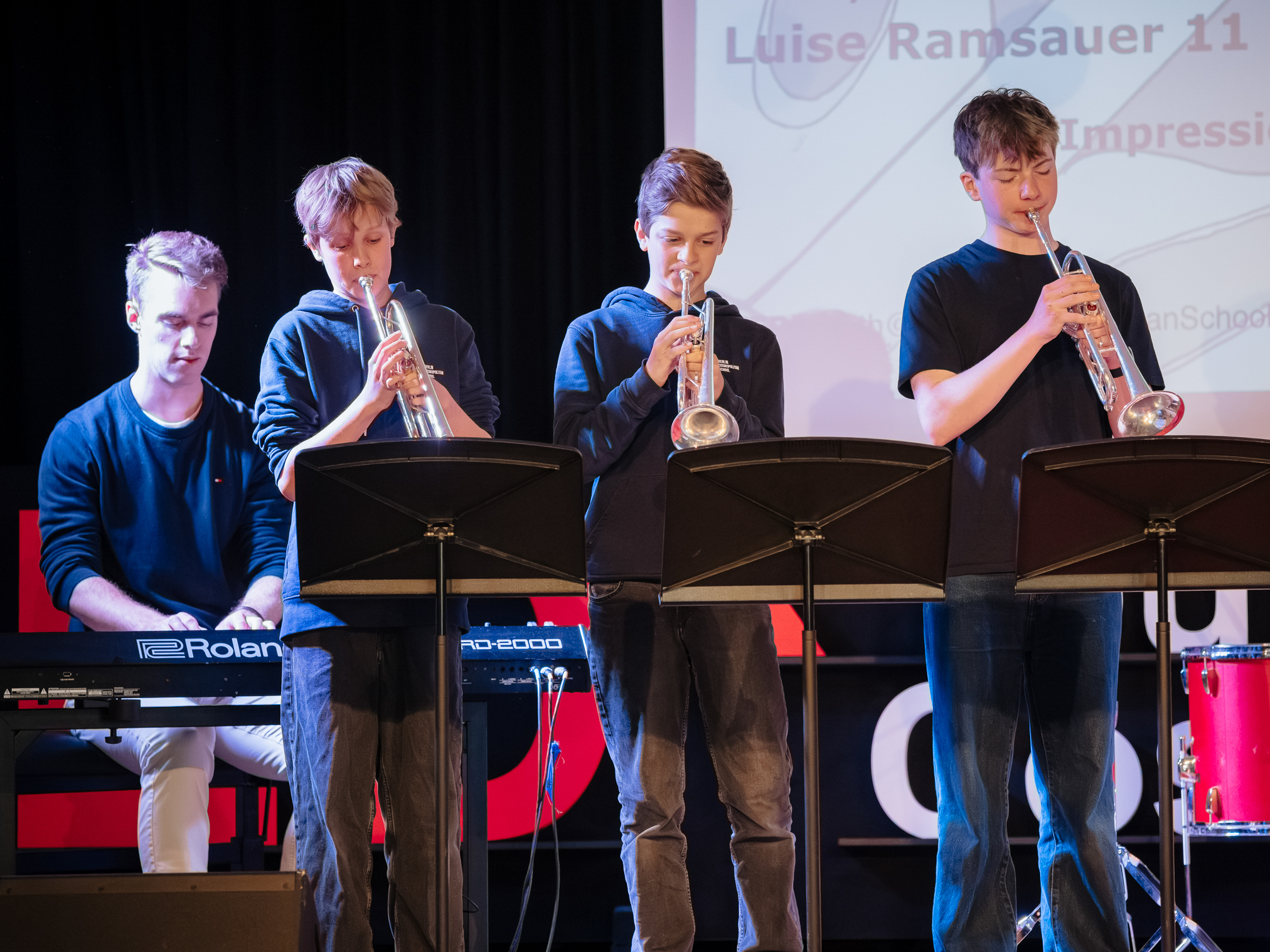 Cosmopolitanschool Berlin Vier Jugendliche treten auf der Bühne auf; einer spielt ein Roland-Keyboard, die anderen drei spielen jeweils eine Trompete. Sie stehen hinter Notenständern, sind leger gekleidet und hinter ihnen wird eine Präsentation projiziert. loading=