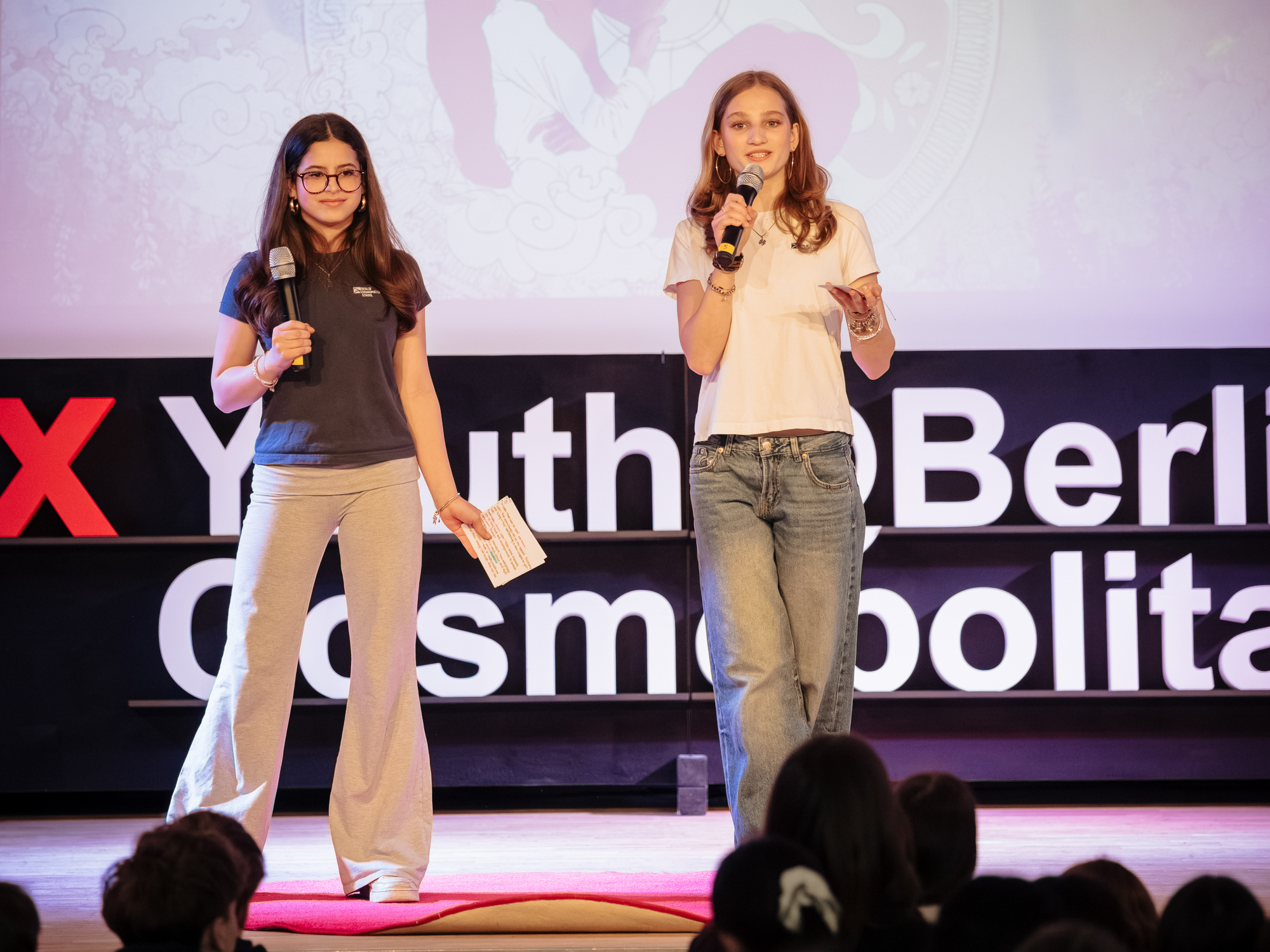 Cosmopolitanschool Berlin Zwei junge Frauen stehen auf der Bühne, halten Mikrofone in der Hand und sprechen zu einem Publikum bei einer TEDxYouth-Veranstaltung in Berlin. Im Hintergrund ist ein TEDxYouthBerlin-Schild zu sehen und im Vordergrund sind Zuhörer zu erkennen. loading=
