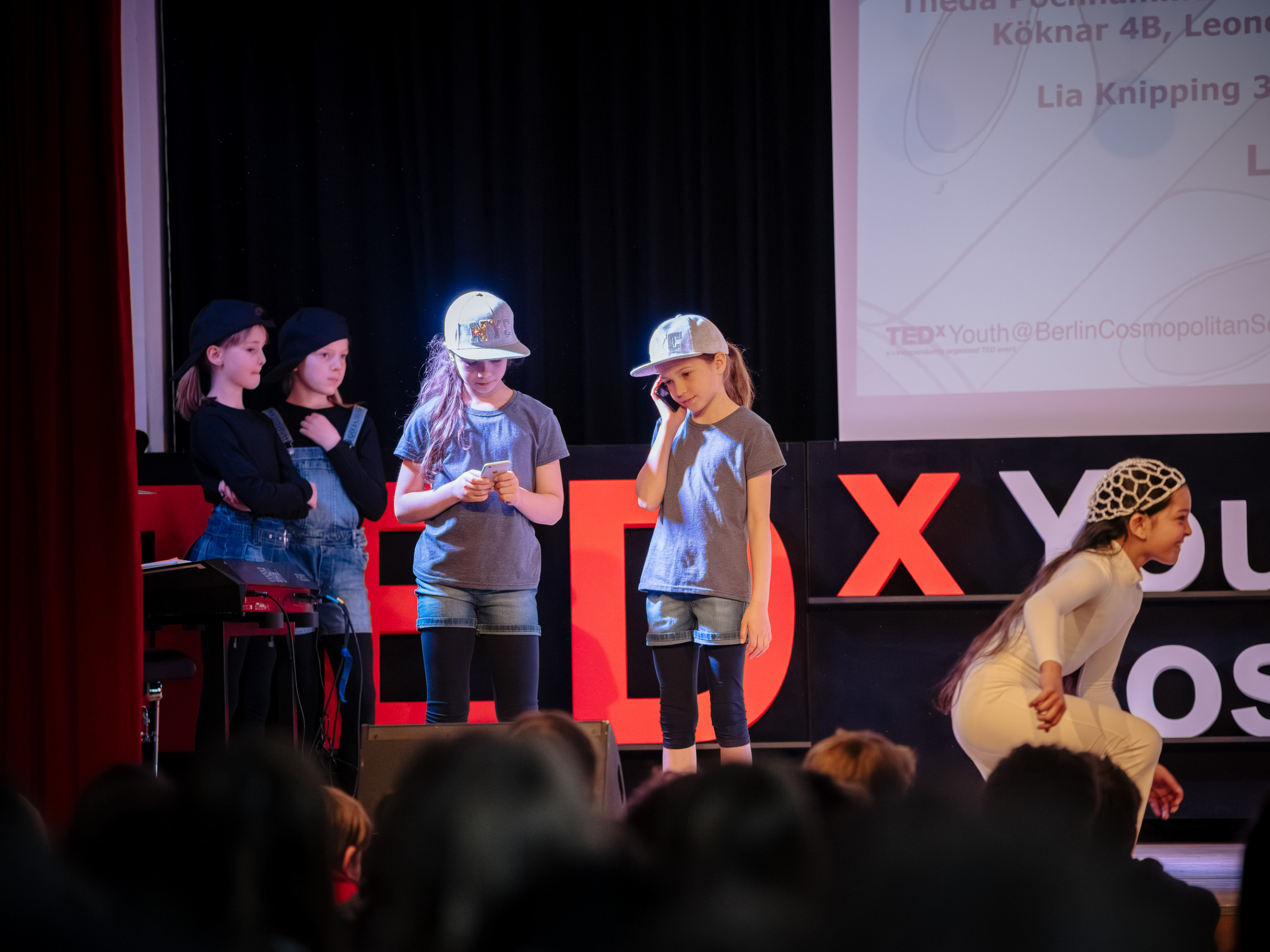 Fünf Kinder auf einer Bühne vor einem TEDx-Youth-Logo, vier stehen nebeneinander, eines ist nach vorne gebeugt, alle tragen lässige Kleidung und Mützen. loading=