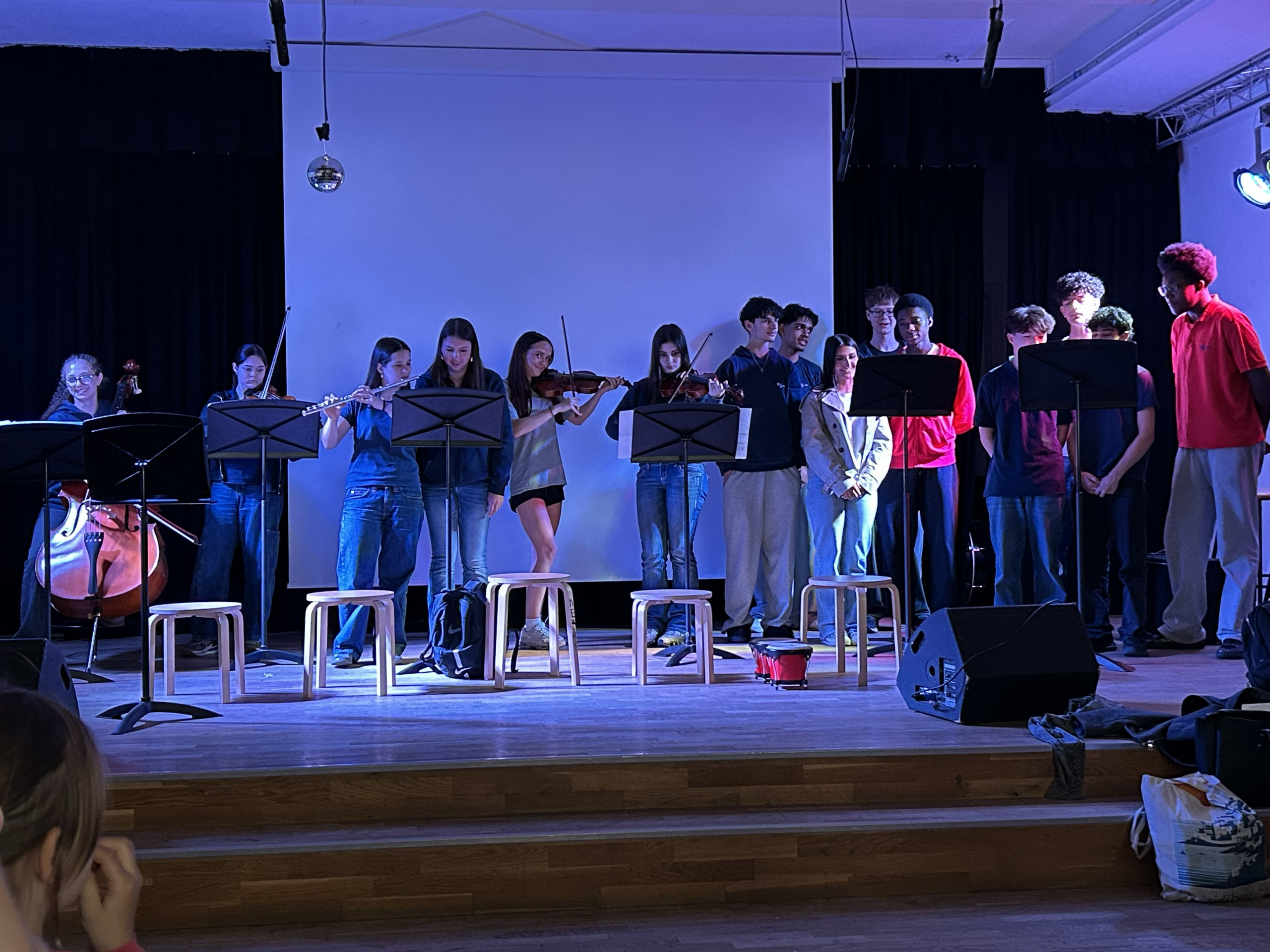Cosmopolitanschool Berlin Eine Gruppe von Schülern steht auf einer Bühne, die meisten spielen Streichinstrumente wie Geige und Cello, während andere nur zuschauen. Vor ihnen stehen Notenständer, hinter ihnen befindet sich eine weiße Leinwand. Die Bühne ist mit blauem Licht beleuchtet.