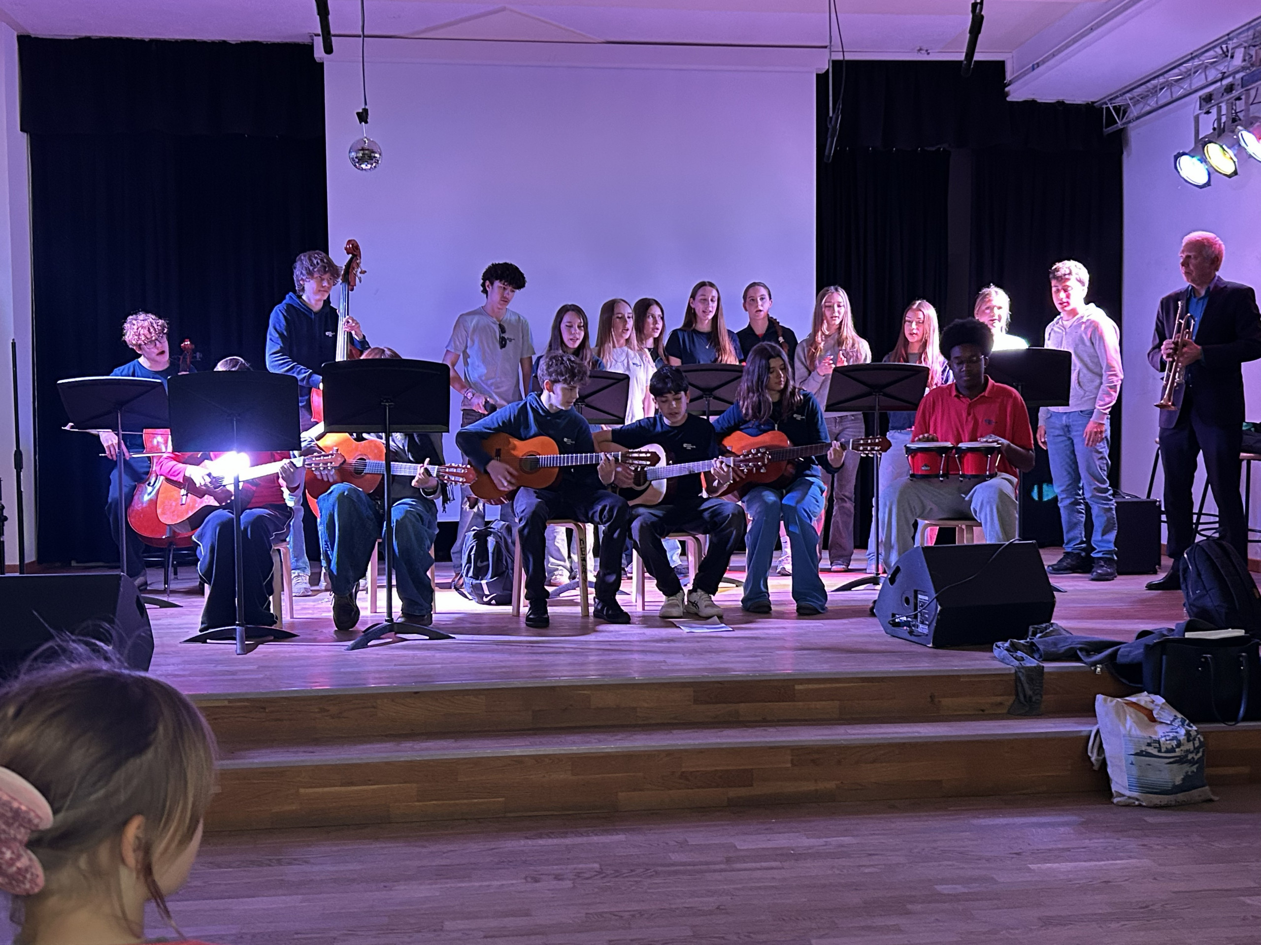 Cosmopolitanschool Berlin Eine Gruppe von Schülern spielt Gitarren, Schlagzeug und singt auf der Bühne, während an den Seiten ein Trompeter und ein Bassist stehen. Bühnenlicht und Mikrofone beleuchten die Interpreten bei einem Schulkonzert. Ein Zuhörer sitzt vorne im Publikum.