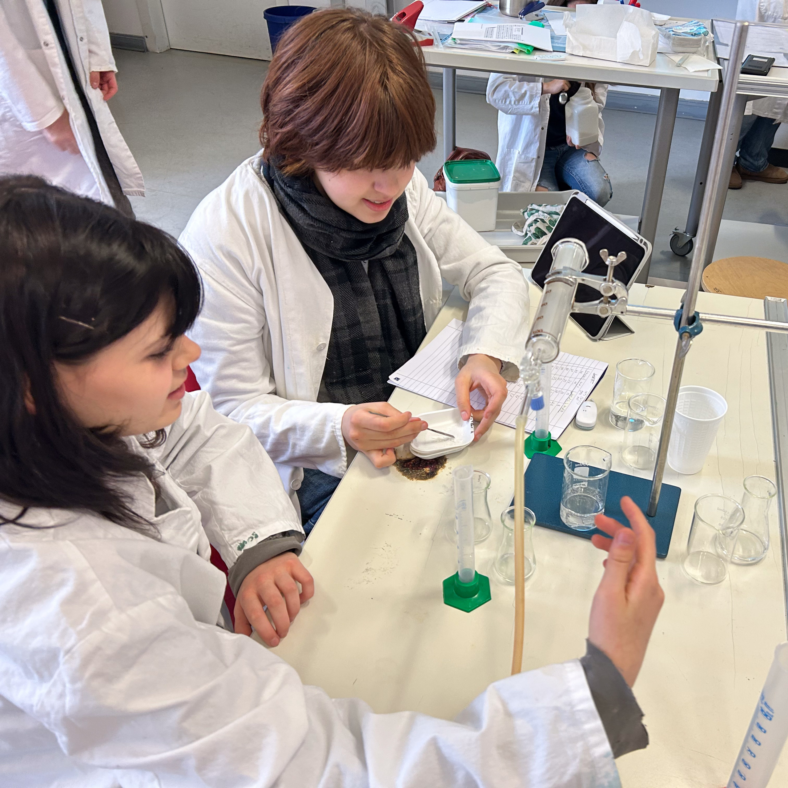 Cosmopolitanschool Berlin Zwei Studenten in weißen Laborkitteln führen an einem Labortisch ein Experiment mit Glaswaren und wissenschaftlichen Geräten durch, während im Hintergrund Notizbücher und andere Studenten zu sehen sind. loading=