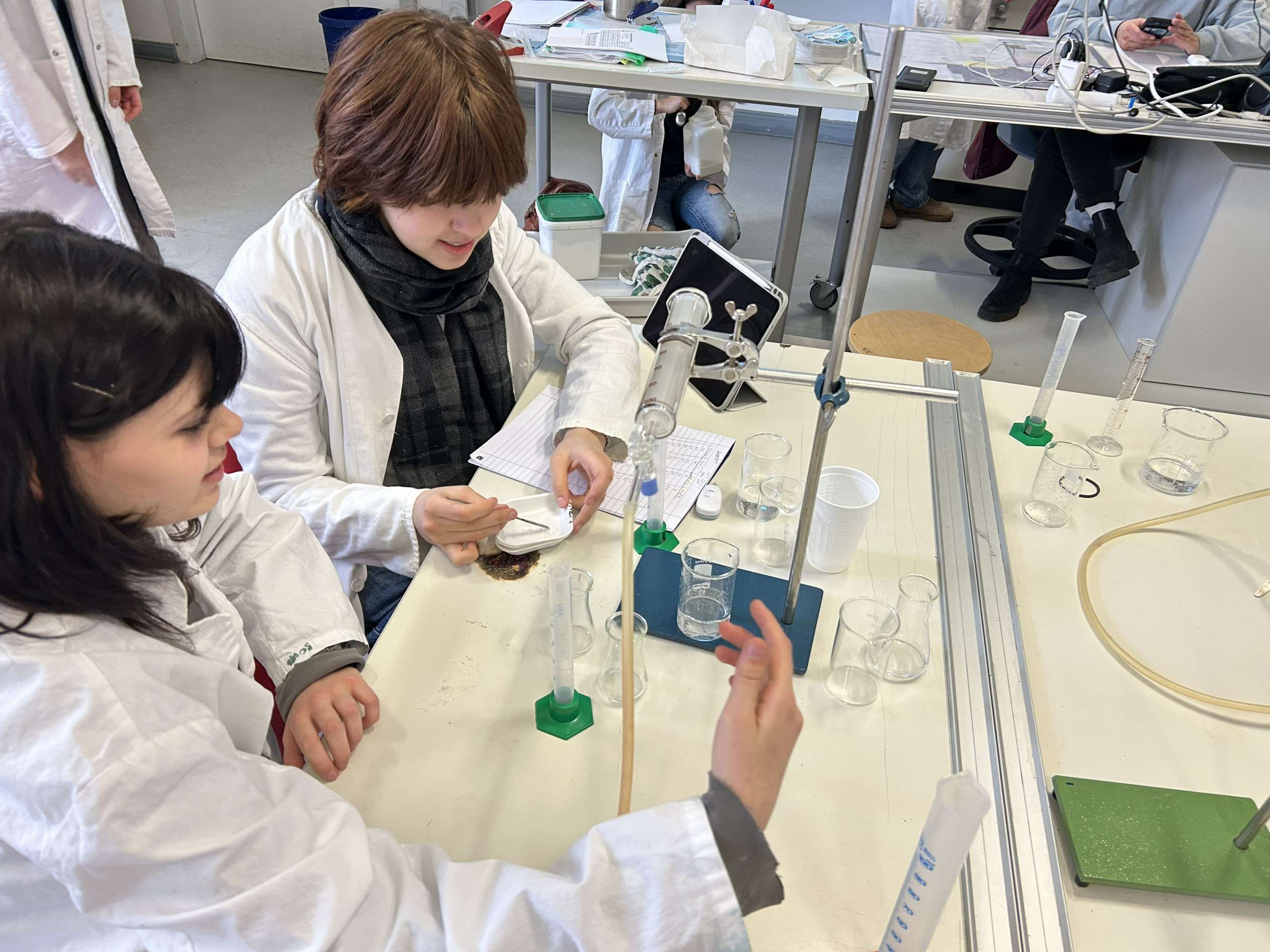 Cosmopolitanschool Berlin Zwei Studenten in Laborkitteln arbeiten gemeinsam an einem Labortisch mit Glaswaren und wissenschaftlichen Geräten und führen ein Experiment durch. Einer schreibt Notizen, während der andere mit einer Petrischale hantiert. Andere Studenten sind im Hintergrund zu sehen. loading=