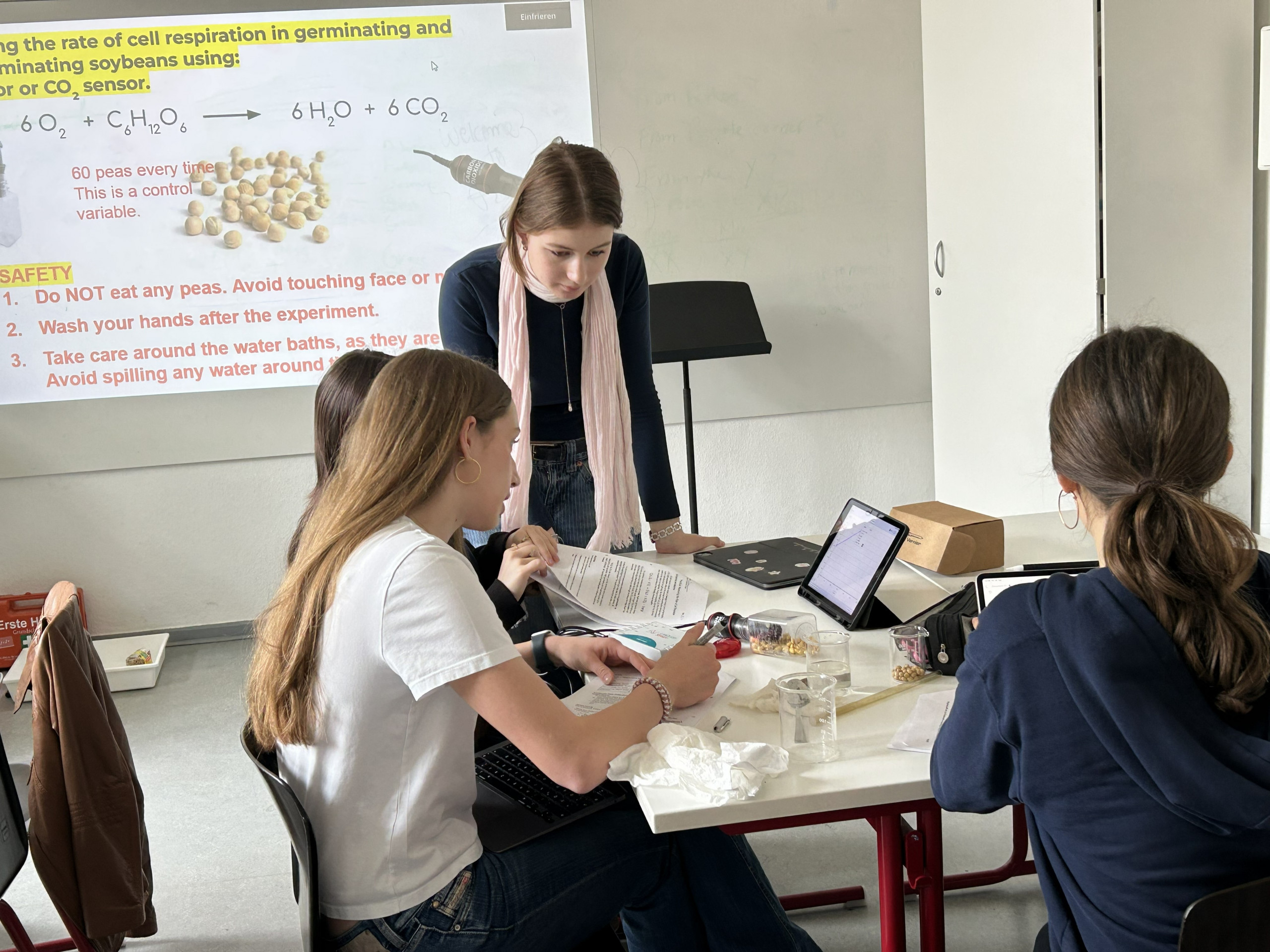 Cosmopolitanschool Berlin Drei Schüler sitzen an einem Tisch und arbeiten mit Arbeitsblättern und Laptops an einem wissenschaftlichen Experiment, während ein Lehrer daneben steht und ihnen hilft. Eine Sicherheitsanweisung und ein Atmungsdiagramm werden auf einen Bildschirm im Hintergrund projiziert. loading=