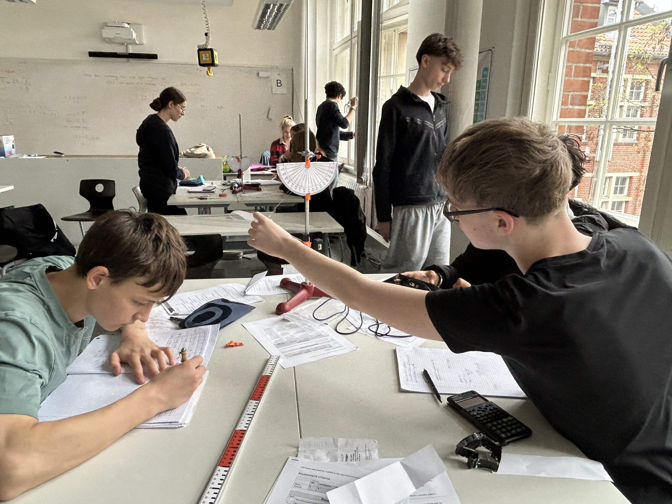 Cosmopolitanschool Berlin Schüler arbeiten an wissenschaftlichen Experimenten und Berechnungen in einem Klassenzimmer. Ein Schüler hält einen Winkelmesser und eine Schnur, während ein anderer Notizen schreibt. Laborgeräte, Papiere und Taschenrechner liegen verstreut auf dem Tisch. Andere Schüler arbeiten im Hintergrund. loading=