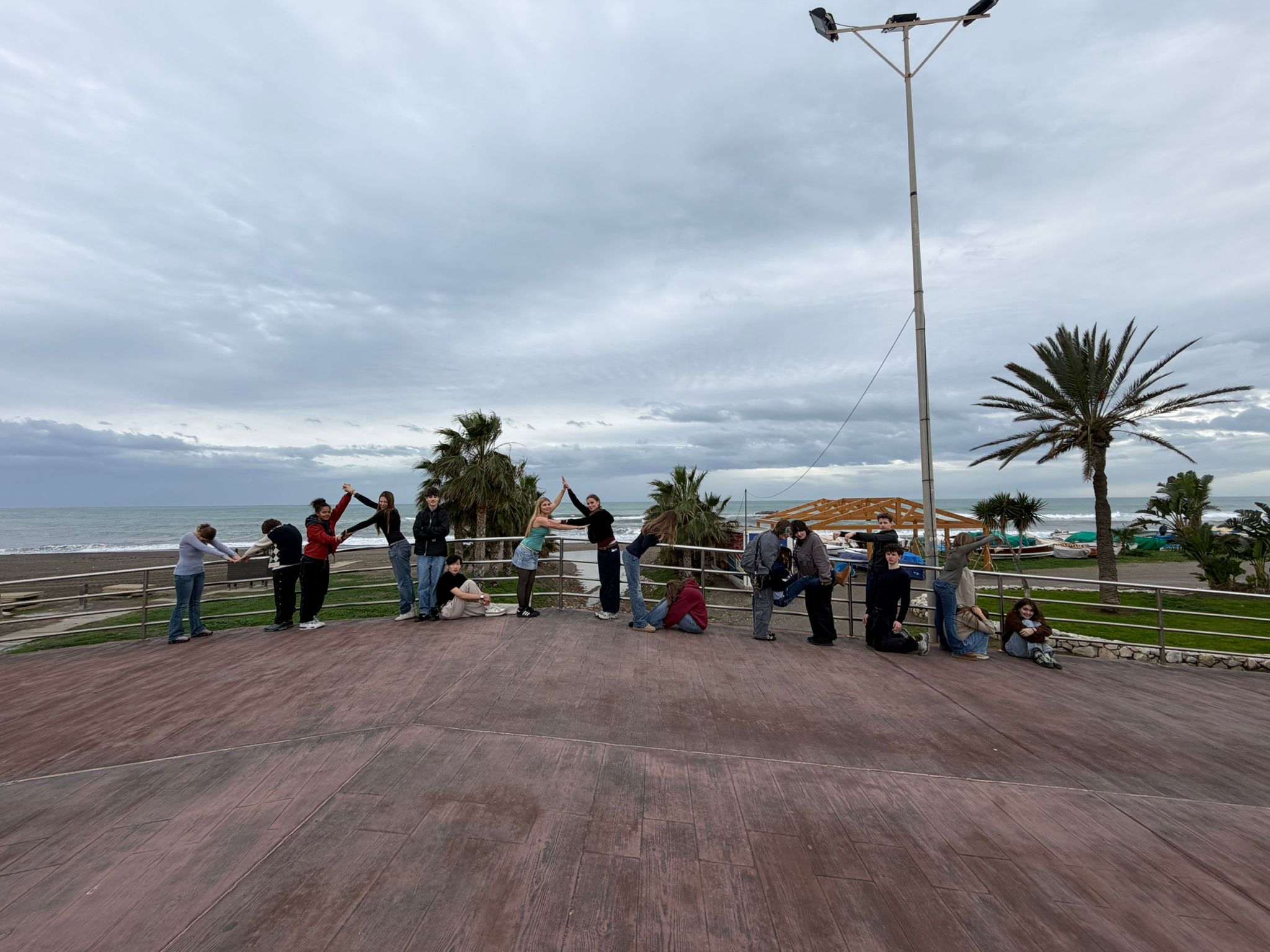 Cosmopolitanschool Berlin Eine Gruppe von Menschen steht und sitzt an einer Strandpromenade, einige heben ihre Arme in spielerischer Pose. Palmen, Gras und ein wolkenverhangener Himmel bilden eine malerische Küstenkulisse. loading=