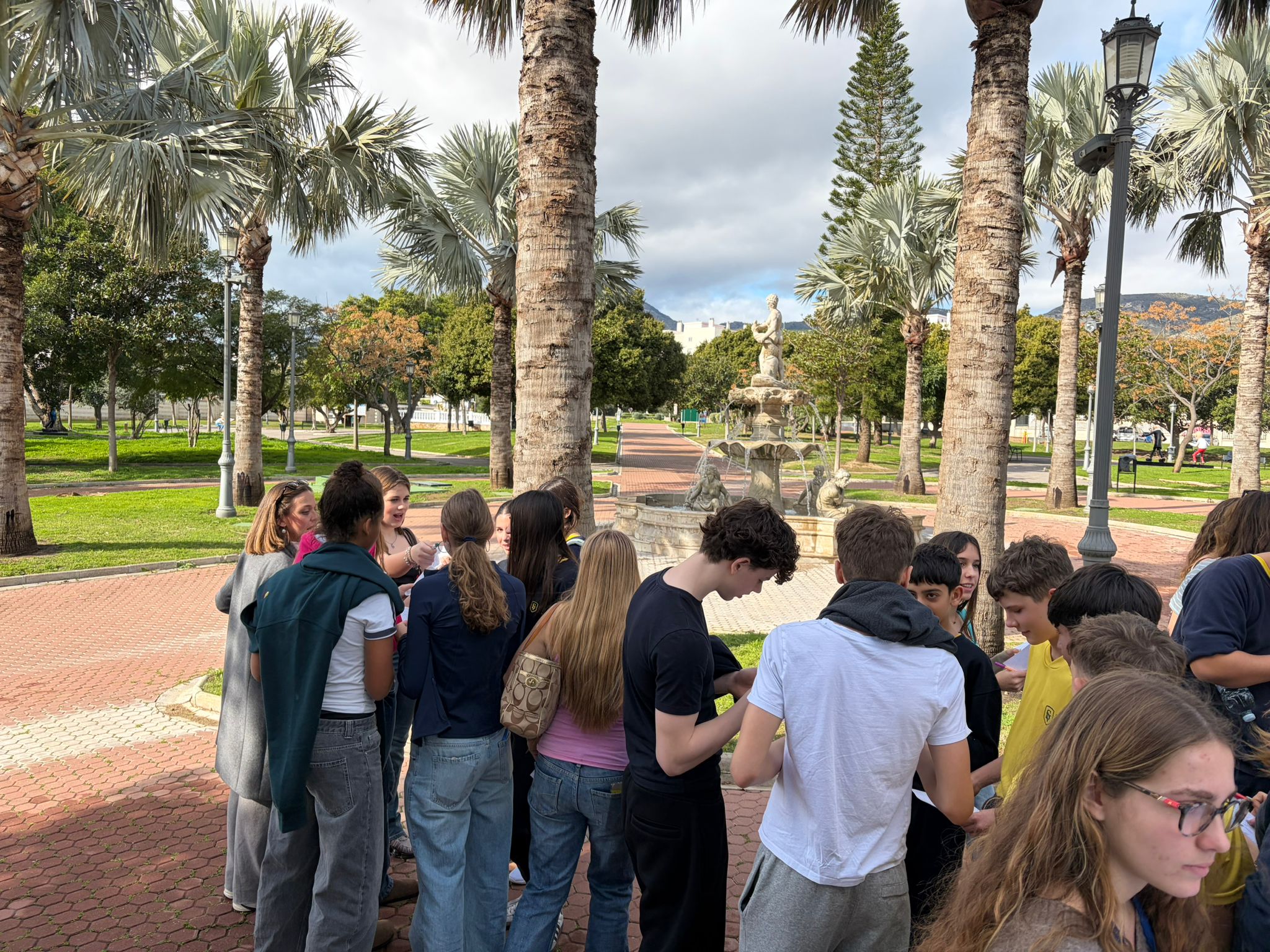 Cosmopolitanschool Berlin Eine Gruppe junger Leute versammelt sich in einem Park in der Nähe hoher Palmen und eines dekorativen Steinbrunnens. Einige plaudern, andere schauen auf Papiere, unter einem teilweise bewölkten Himmel. Der Park ist grün und gut gepflegt. loading=