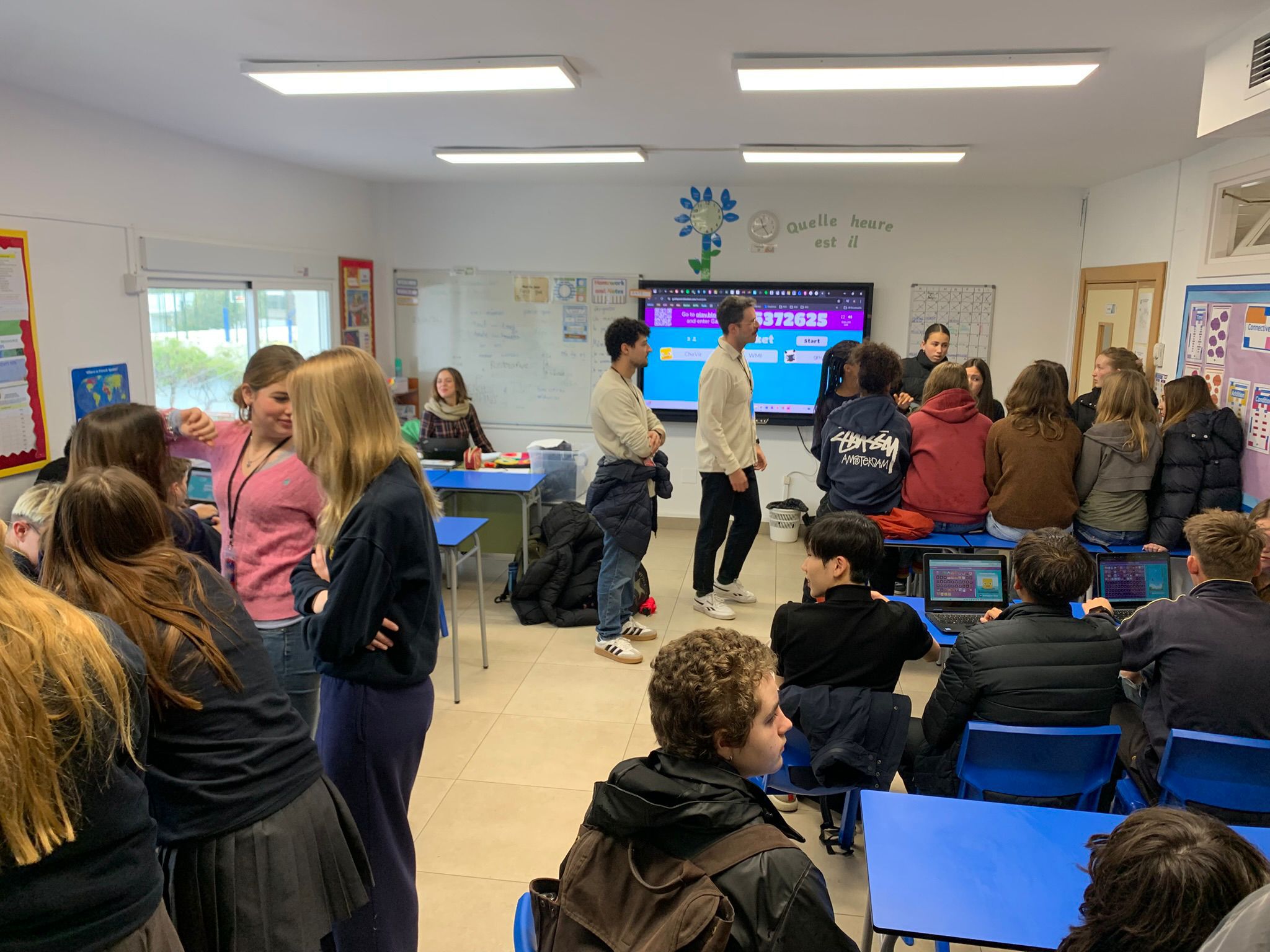 Cosmopolitanschool Berlin Ein Klassenzimmer voller Schüler, von denen einige an blauen Pulten sitzen und andere in Gruppen stehen und sich unterhalten. Ein Lehrer steht vorne neben einem großen Bildschirm, auf dem bunte Informationen angezeigt werden. Die Atmosphäre wirkt lebendig und interaktiv. loading=