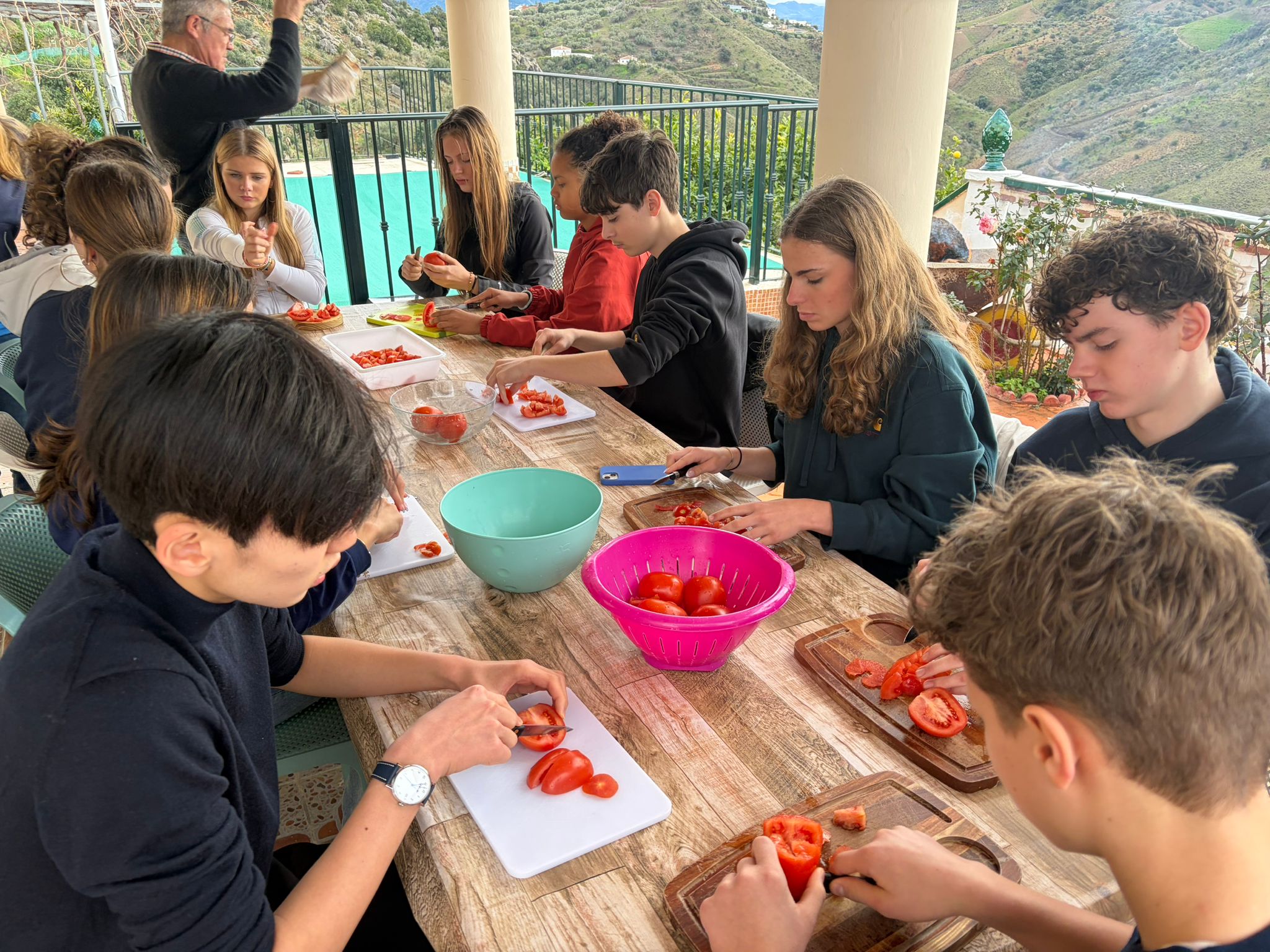 Cosmopolitanschool Berlin Eine Gruppe Jugendlicher sitzt um einen Holztisch im Freien und schneidet mit Messern und Schneidebrettern Tomaten. Große Schüsseln mit geschnittenen Tomaten stehen auf dem Tisch. Im Hintergrund sind Berge und Grünflächen zu sehen. loading=