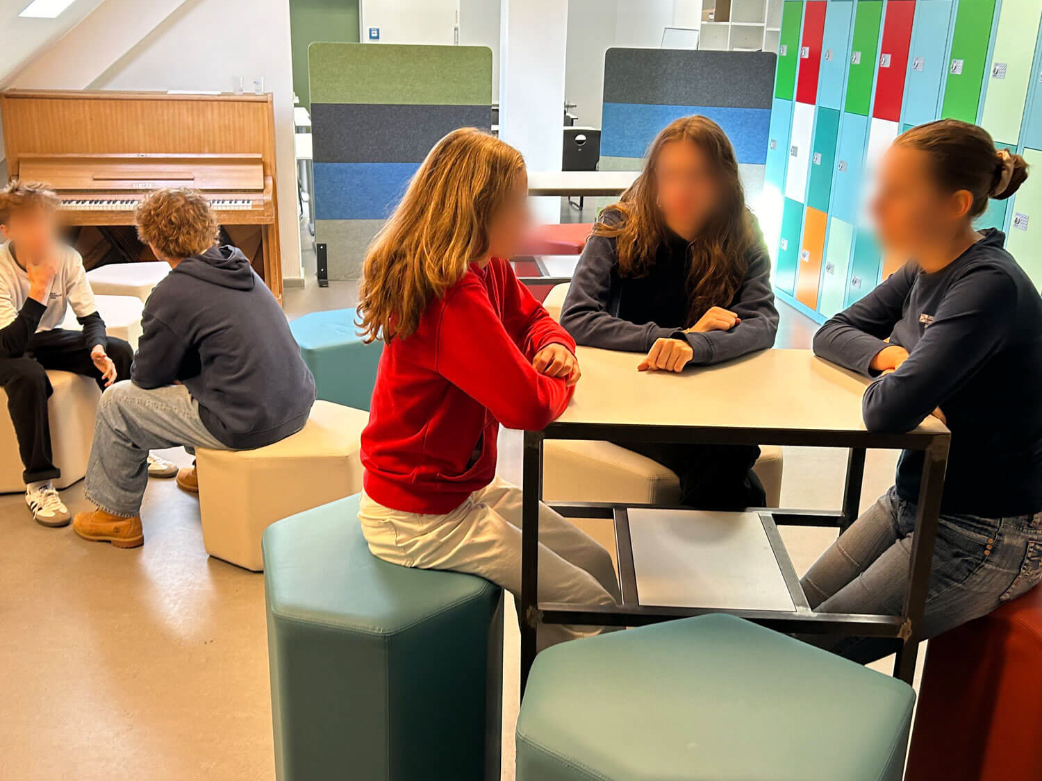 Cosmopolitanschool Berlin Mehrere Schüler sitzen in Gruppen in einem hell erleuchteten Klassenzimmer mit bunten Spinden, modernen Möbeln und einem Klavier im Hintergrund und unterhalten sich. Die Gesichter sind zum Schutz der Privatsphäre unscharf.