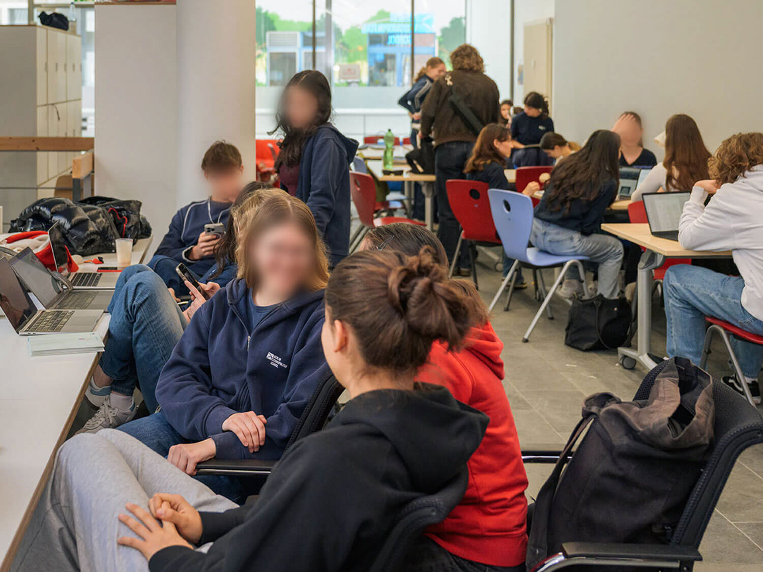 Cosmopolitanschool Berlin Eine Gruppe von Schülern sitzt in einem hellen Klassenzimmer mit großen Fenstern und unterhält sich. Einige arbeiten mit Laptops, andere chatten oder schauen auf ihr Handy. Der Raum ist mit bunten Stühlen und Rucksäcken ausgestattet. Die Gesichter sind unscharf, um die Privatsphäre zu wahren.