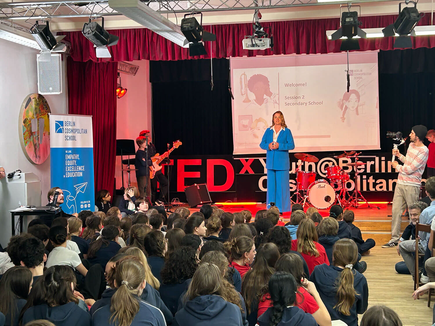 Cosmopolitanschool Berlin Eine Frau in einem blauen Anzug spricht auf der Bühne einer TEDx-Jugendveranstaltung in einer Schulaula. Auf dem Boden sitzen Schüler, Musiker, ein Kameramann und ein Banner der Berlin Cosmopolitan School ist zu sehen.
