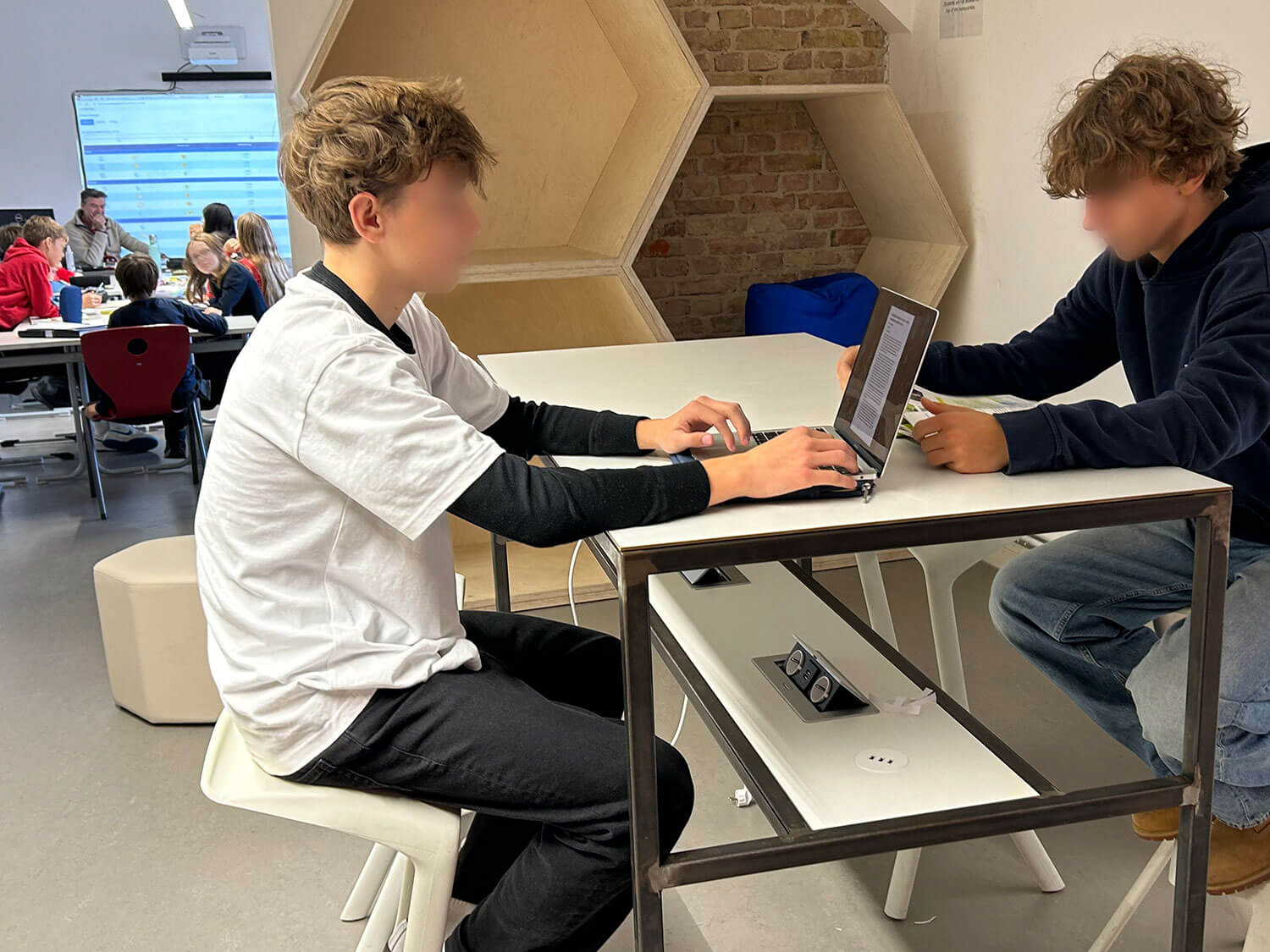 Cosmopolitanschool Berlin Zwei Jugendliche sitzen an einem Stehtisch und arbeiten an Laptops in einem modernen Klassenzimmer. Im Hintergrund sind andere Schüler und ein Lehrer zu sehen. Sechseckige Regale und eine Backsteinmauer sind Teil der Einrichtung.
