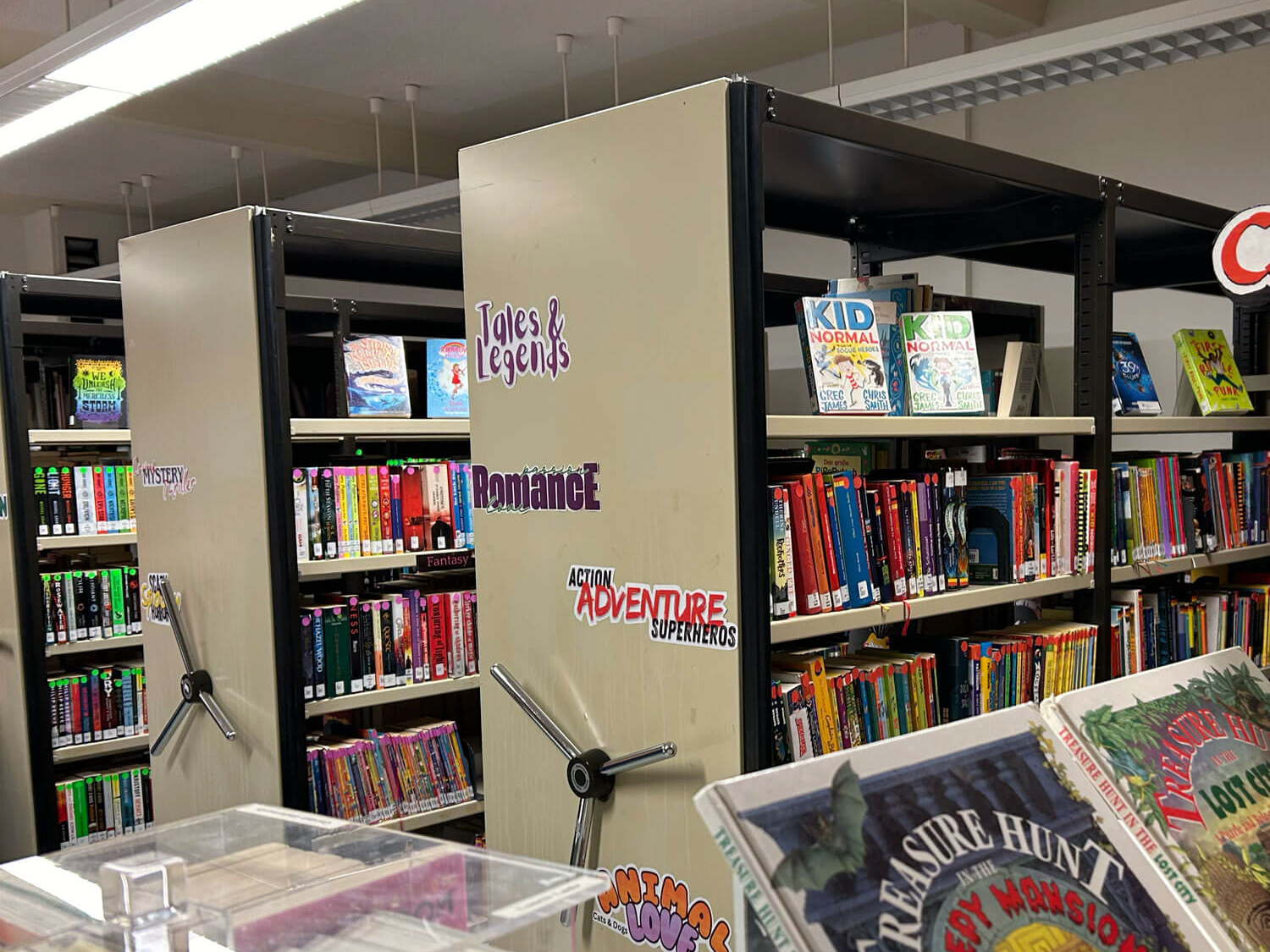 Cosmopolitanschool Berlin Eine Bibliothek mit Regalen mit farbenfrohen Kinderbüchern, beschriftet mit Rubriken wie "Märchen und Legenden", "Romantik", "Action-Abenteuer-Superhelden" und "Tierwelt". Im Vorder- und Hintergrund sind Buchumschläge zu sehen.