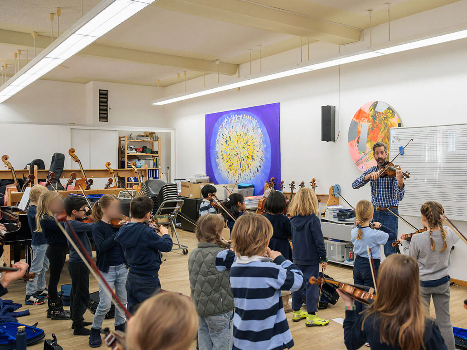 Cosmopolitanschool Berlin Eine Gruppe von Kindern spielt in einem Klassenzimmer Geige, während sie einem erwachsenen Lehrer gegenübersitzen. Der Raum ist hell beleuchtet, hat Notenständer und bunte Kunstwerke an den Wänden.