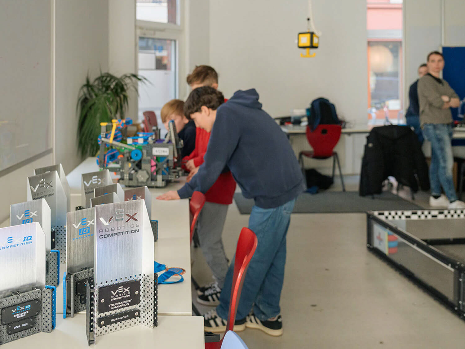 Cosmopolitanschool Berlin Eine Gruppe von Schülerinnen und Schülern arbeitet an einem Tisch in einem hellen Klassenzimmer an Robotikprojekten. Im Vordergrund sind die Trophäen des VEX-Wettbewerbs zu sehen, rechts eine Roboterarena.