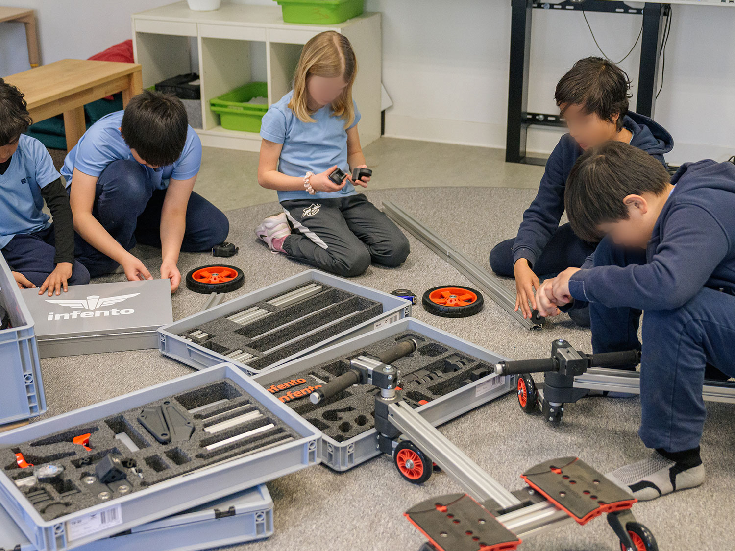 Cosmopolitanschool Berlin Fünf Kinder sitzen auf dem Boden und bauen einen Bausatz mit Metallstäben, Rädern und Teilen zusammen, umgeben von offenen Werkzeugkoffern. Sie wirken konzentriert und arbeiten gemeinsam in einem hellen Klassenzimmer. Die Gesichter sind zum Schutz der Privatsphäre unscharf.