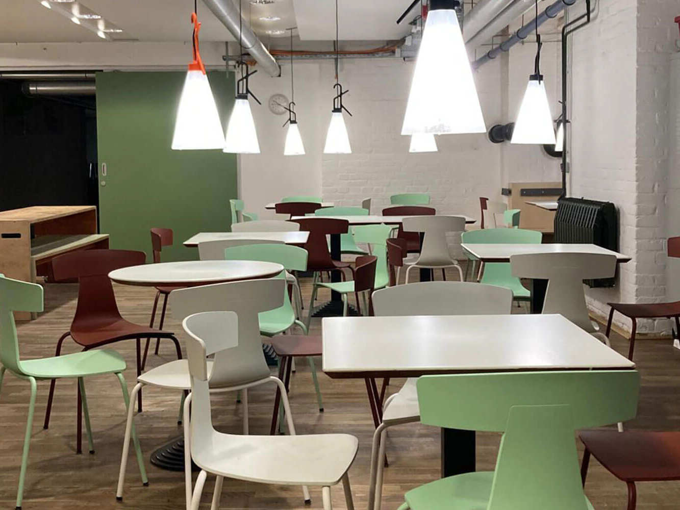 Cosmopolitanschool Berlin Ein modernes Café mit weißen Wänden, Holzböden und verschiedenen quadratischen und runden Tischen, umgeben von mintgrünen und roten Stühlen. Mehrere große Pendelleuchten hängen von der Decke und erhellen den leeren Raum.