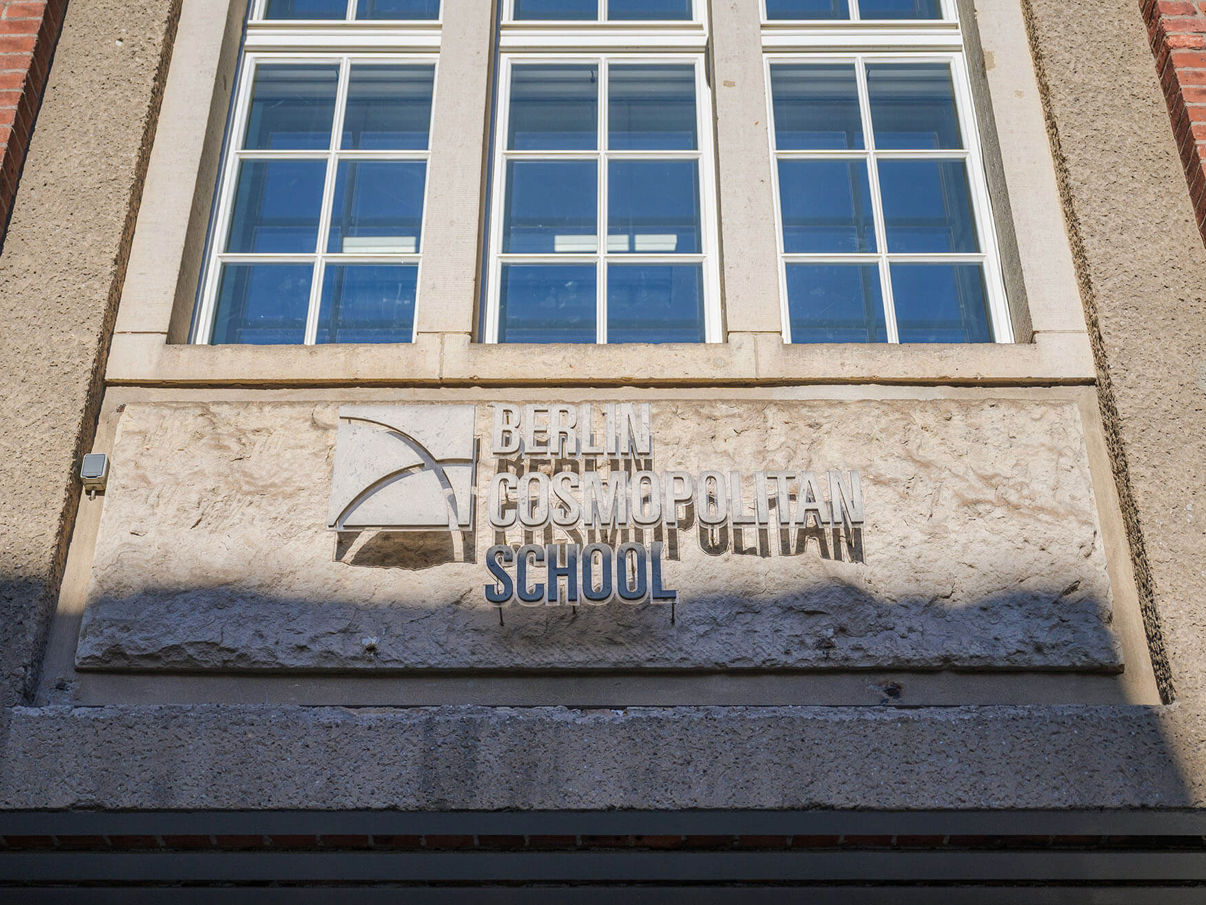 Cosmopolitanschool Berlin Ein steinernes Schild an der Außenseite eines Gebäudes trägt die Aufschrift Berlin Cosmopolitan School unter einer Reihe von großen Fenstern. Das Schild enthält ein minimalistisches Logo mit einer geschwungenen Linie und einem quadratischen Design. loading=