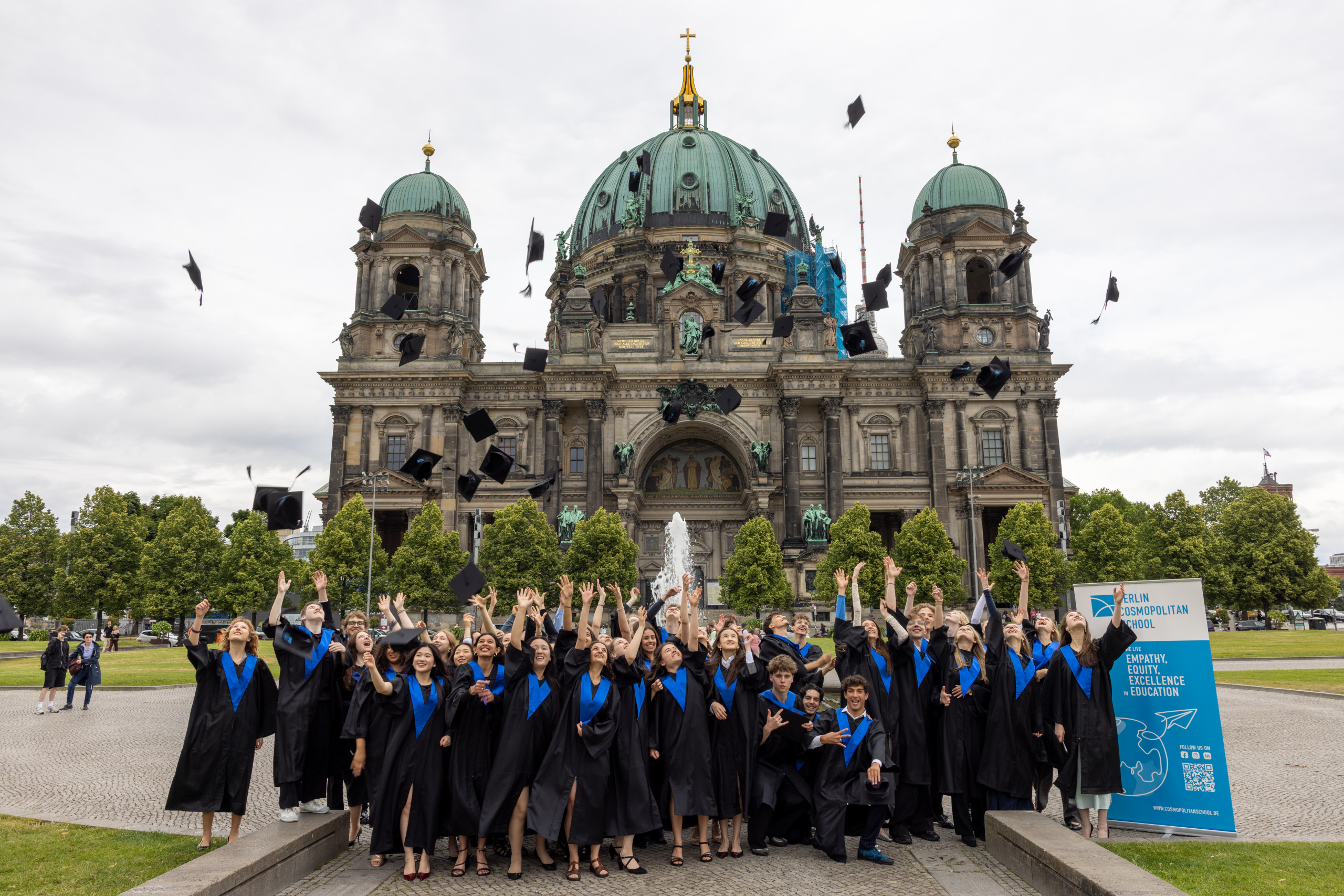 Gruppe von Absolventen in schwarzen Talaren mit blauen Schärpen wirft Hüte vor einer historischen Kuppelkirche in die Luft loading=