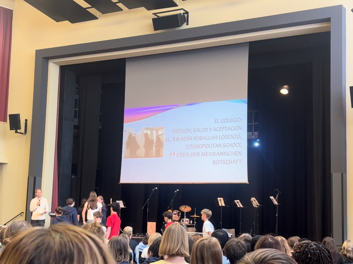 Bühne mit mehreren Personen vor einer Leinwand, auf der ein Text in spanischer Sprache projiziert wird, im Hintergrund Musikinstrumente und Publikum im Vordergrund loading=