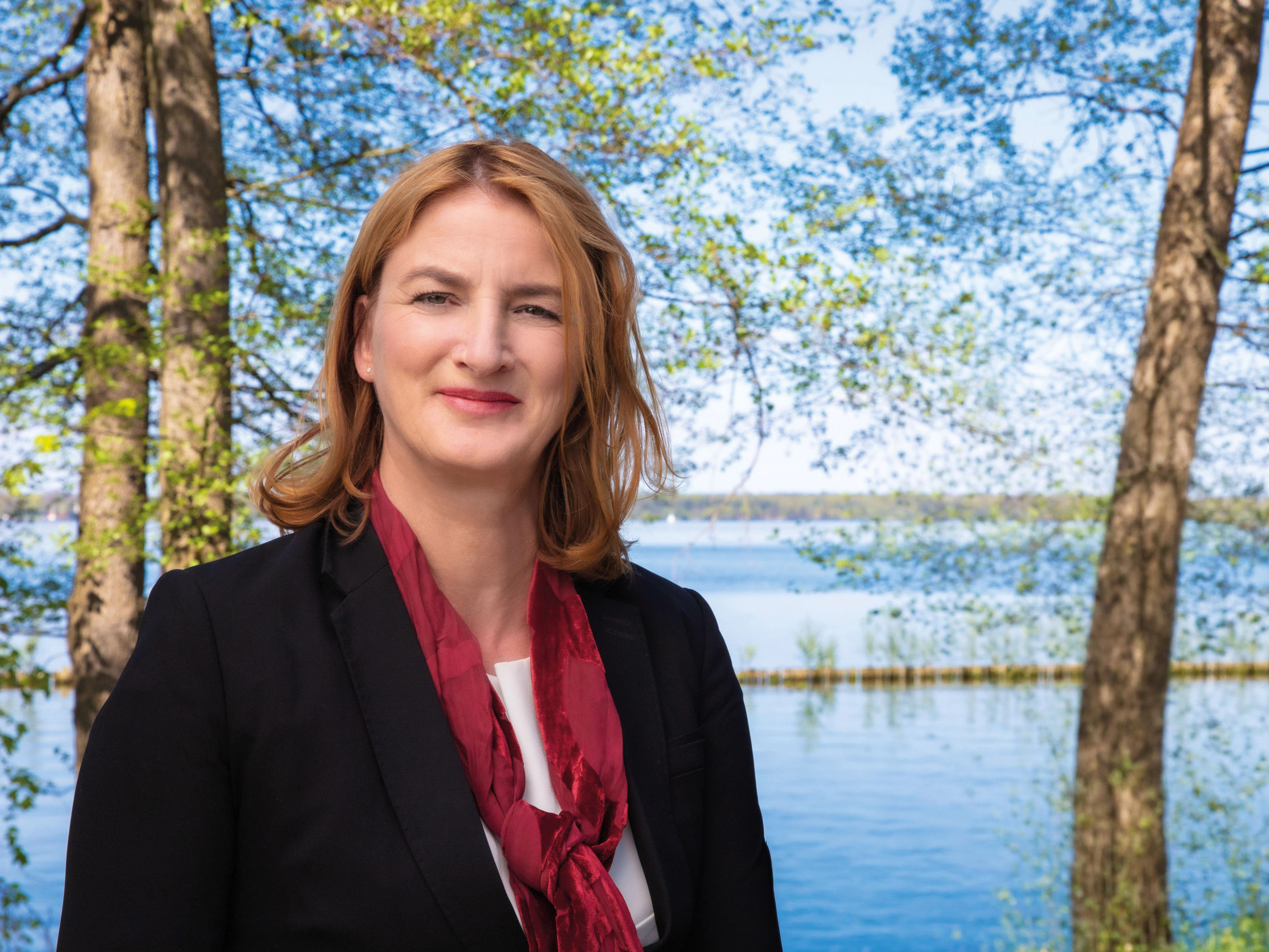 Frau mit schulterlangen blonden Haaren trägt schwarzen Blazer und roten Schal vor einem See mit Bäumen im Hintergrund loading=