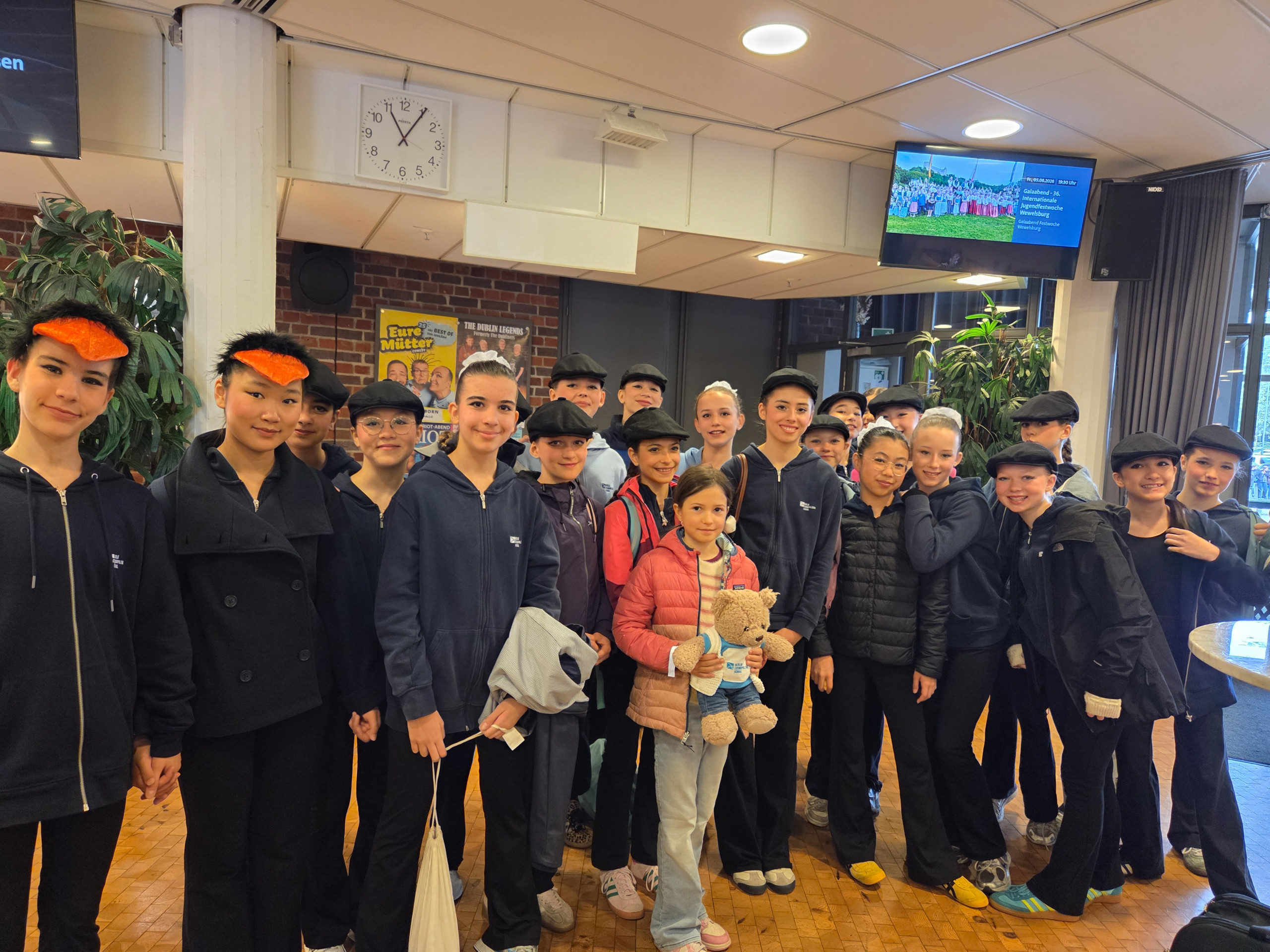 Gruppe von Jugendlichen und Kindern im Foyer vor einer Wand mit Uhr und Monitoren, einige tragen Mützen und eine Person hält zwei Stofftiere loading=