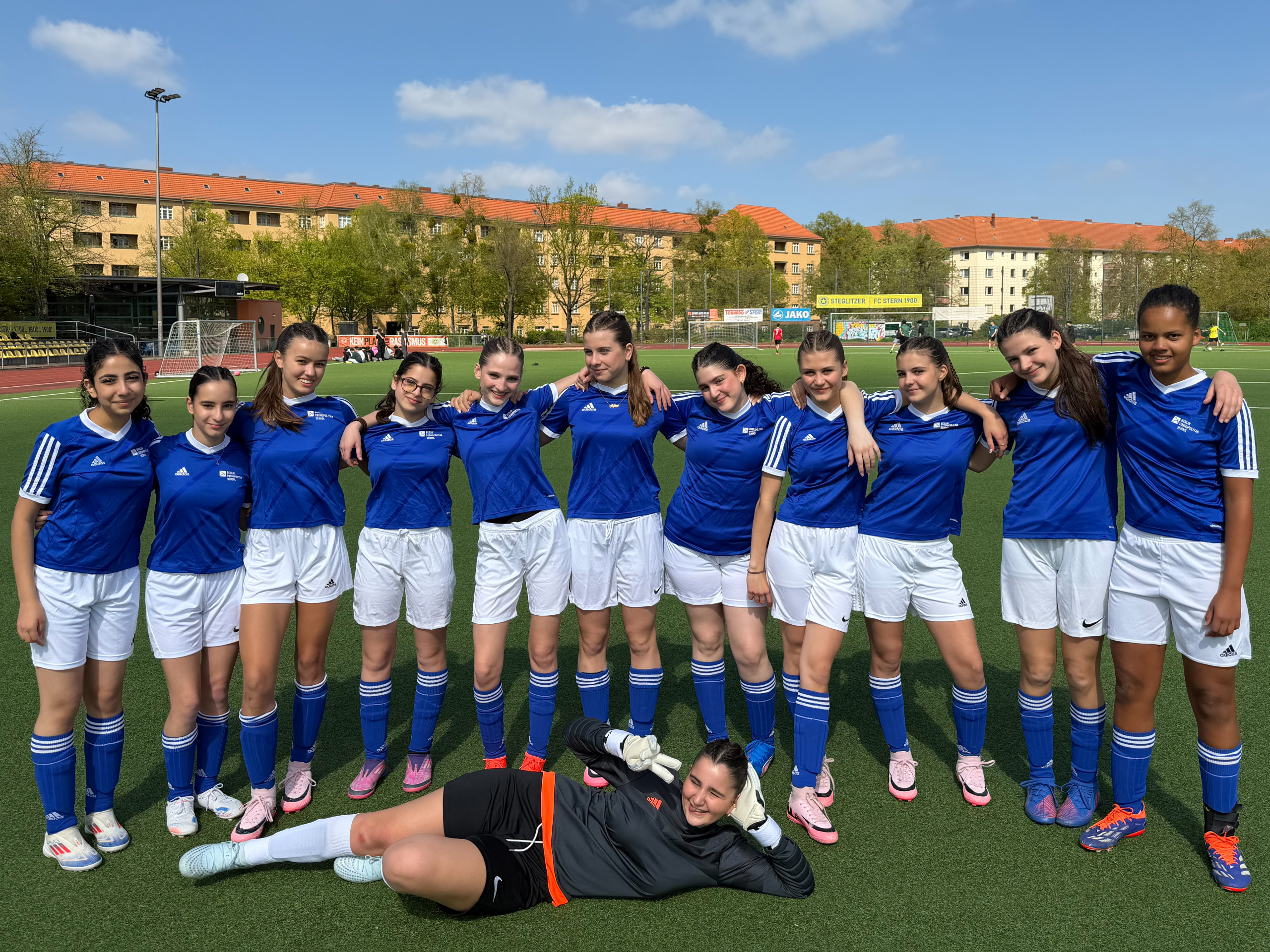Cosmopolitanschool Berlin Elf Fußballspielerinnen in blauen Trikots und weißen Hosen stehen auf einem Fußballfeld, eine liegt vorne auf dem Boden loading=