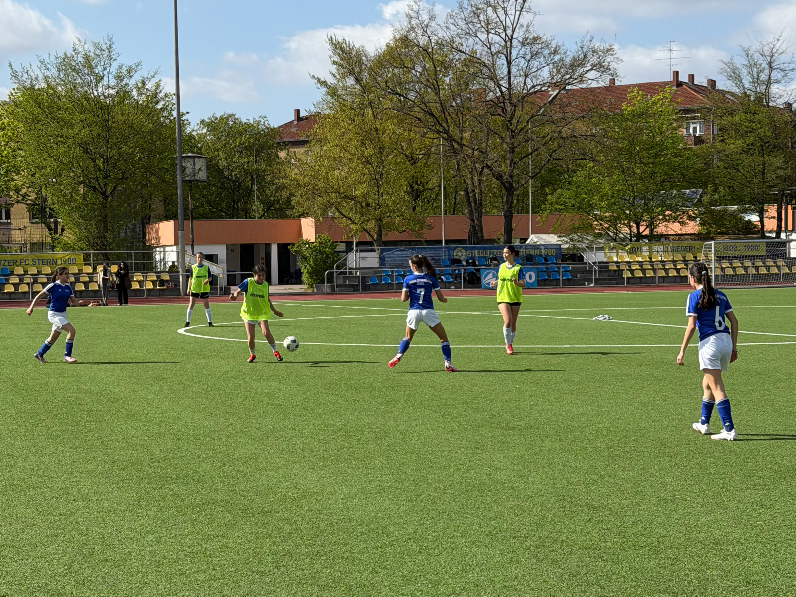 Cosmopolitanschool Berlin Fußballspiel mit Jugendlichen auf einem Kunstrasenplatz der Cosmopolitanschool Berlin, einige Spieler tragen blaue Trikots, andere gelbe Leibchen, im Hintergrund Bäume und ein Gebäude. loading=