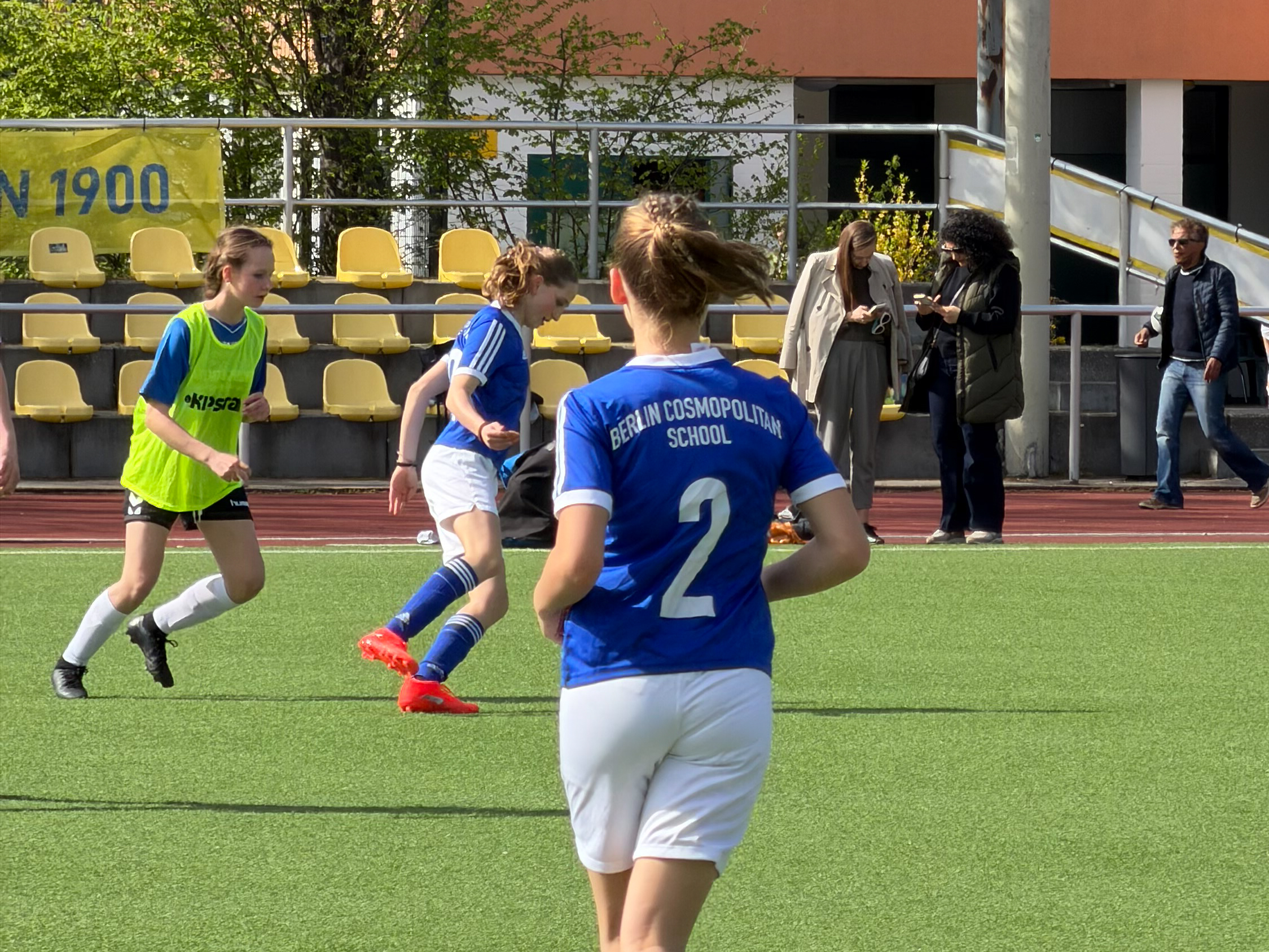 Cosmopolitanschool Berlin Drei Mädchen spielen Fußball auf einem Spielfeld, zwei tragen blaue Trikots, eines ein gelbes Leibchen, im Hintergrund stehen drei Erwachsene an der Seitenlinie loading=