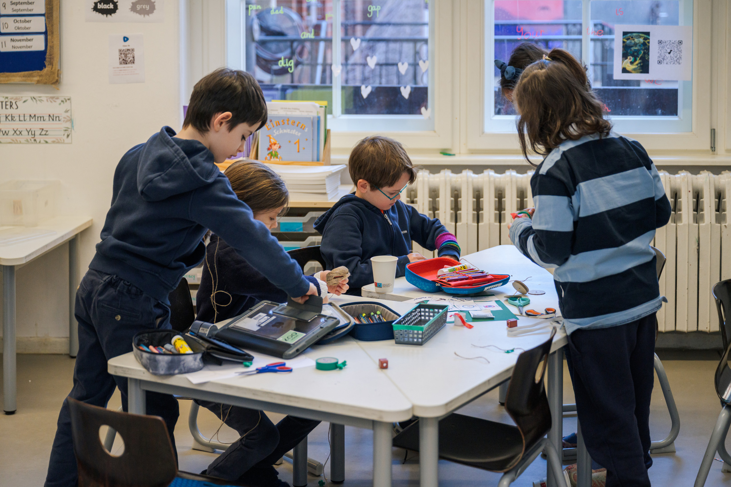 Cosmopolitanschool Berlin Vier Kinder stehen und sitzen um einen Tisch mit Schulmaterialien in einem Klassenraum mit Fenstern und Heizkörpern loading=