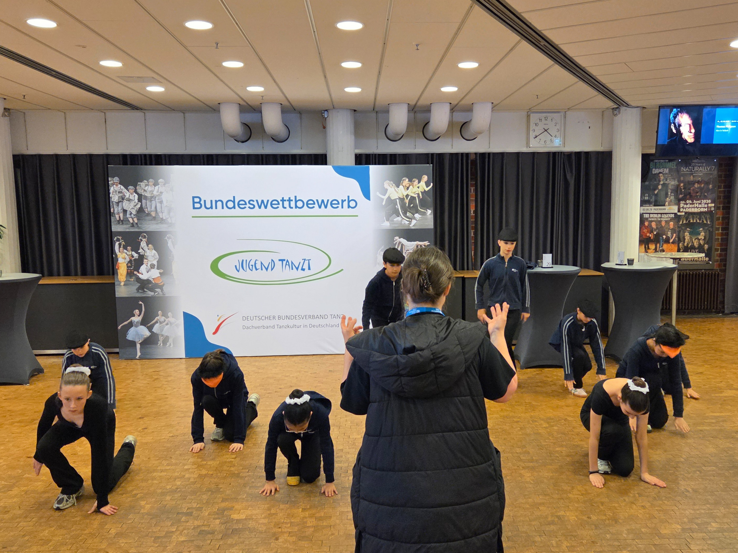 Eine Gruppe Jugendlicher in schwarzer Sportkleidung probt eine Tanzchoreografie vor einer Frau mit schwarzer Weste im Foyer mit einem Banner des Bundeswettbewerbs Jugend tanzt im Hintergrund loading=
