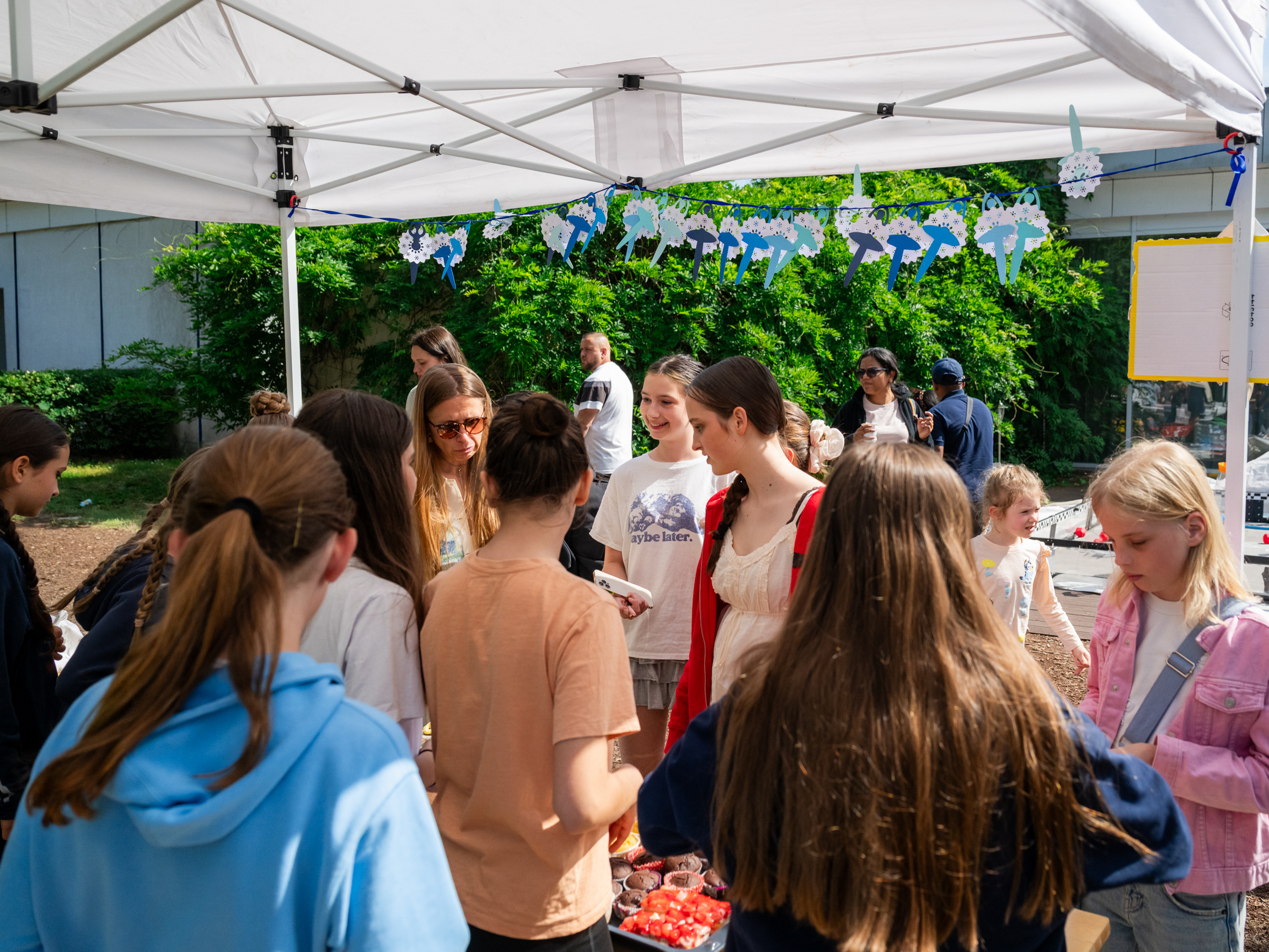 Gruppe von Jugendlichen unter einem weißen Pavillon mit blauen Papiergirlanden, die sich unterhalten und um einen Tisch mit Lebensmitteln versammeln loading=
