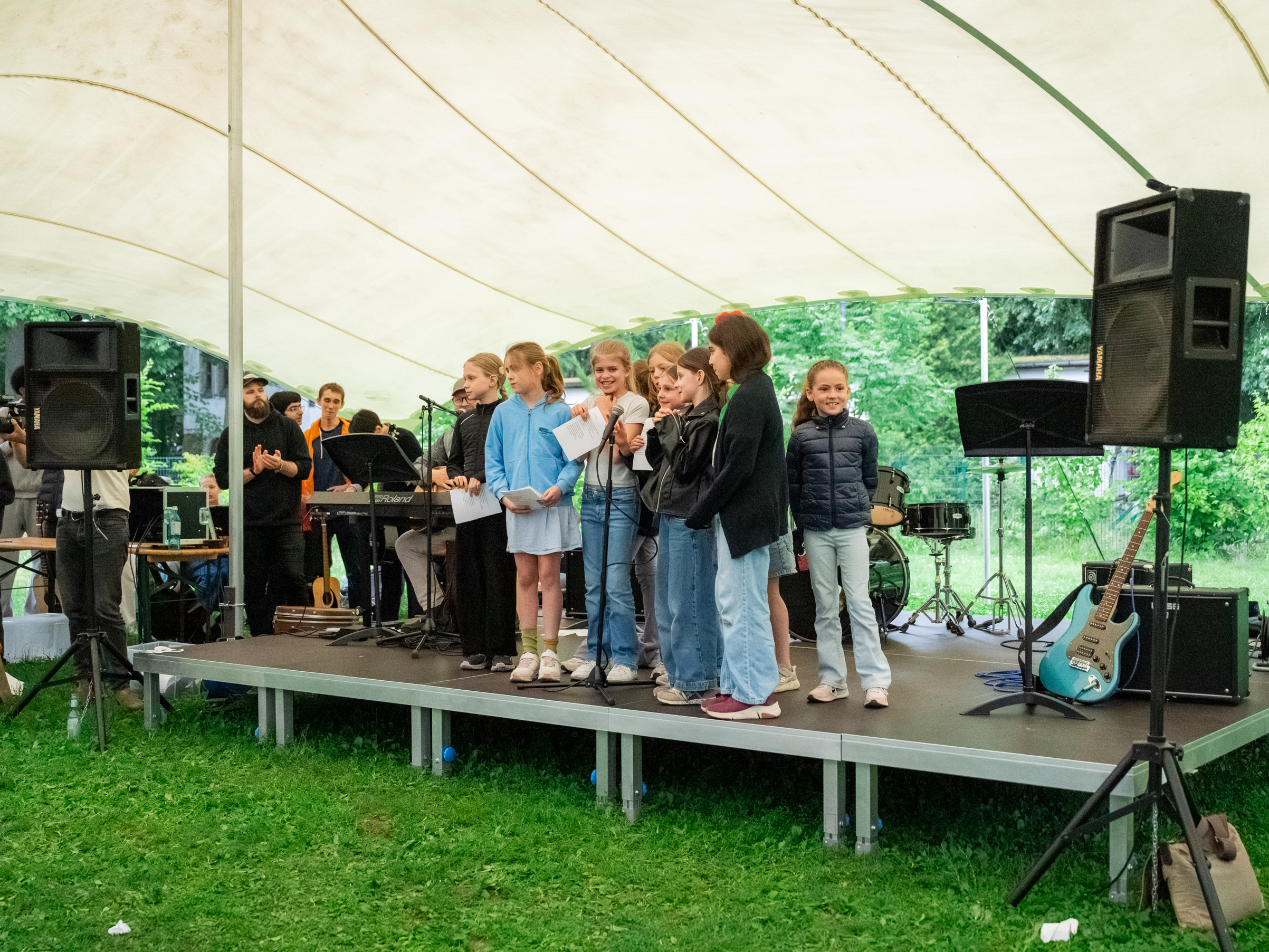 Gruppe von Kindern steht auf einer Bühne unter einem weißen Zeltdach, umgeben von Musikinstrumenten und Lautsprechern loading=