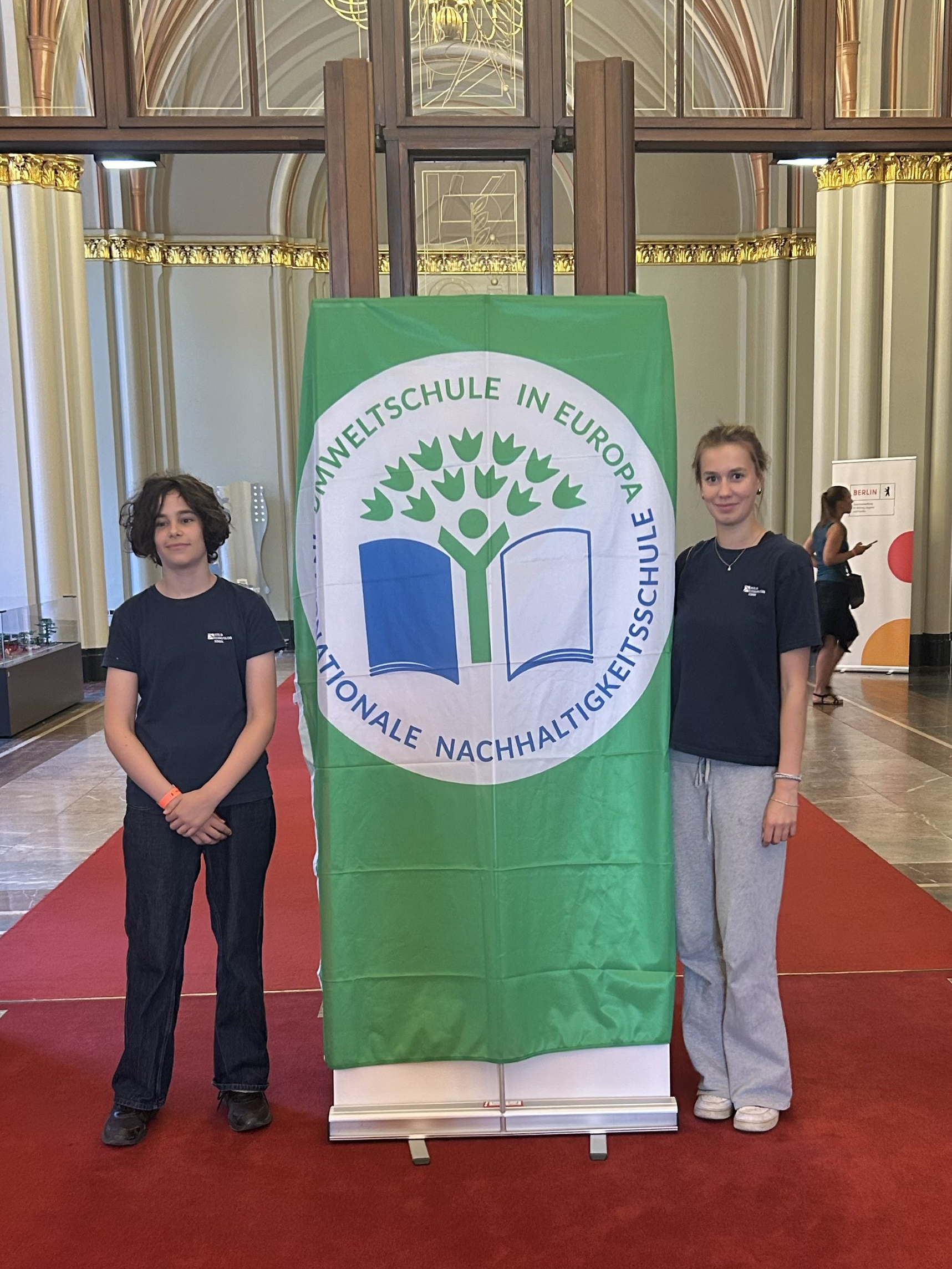 Zwei Personen stehen links und rechts neben einem grünen Banner mit dem Logo der Umweltschule in Europa und dem Text 'Umweltschule in Europa Nationale Nachhaltigkeitsschule' in einem großen, hellen Raum mit hohen Fenstern und rotem Teppich loading=