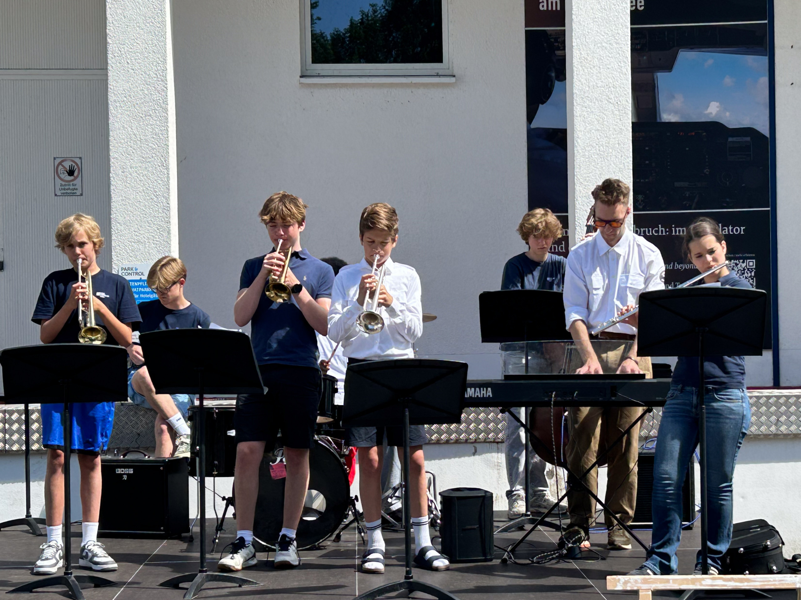 Gruppe von Jugendlichen spielt Trompeten und Keyboard vor einem weißen Gebäude mit Fenster und Plakat loading=