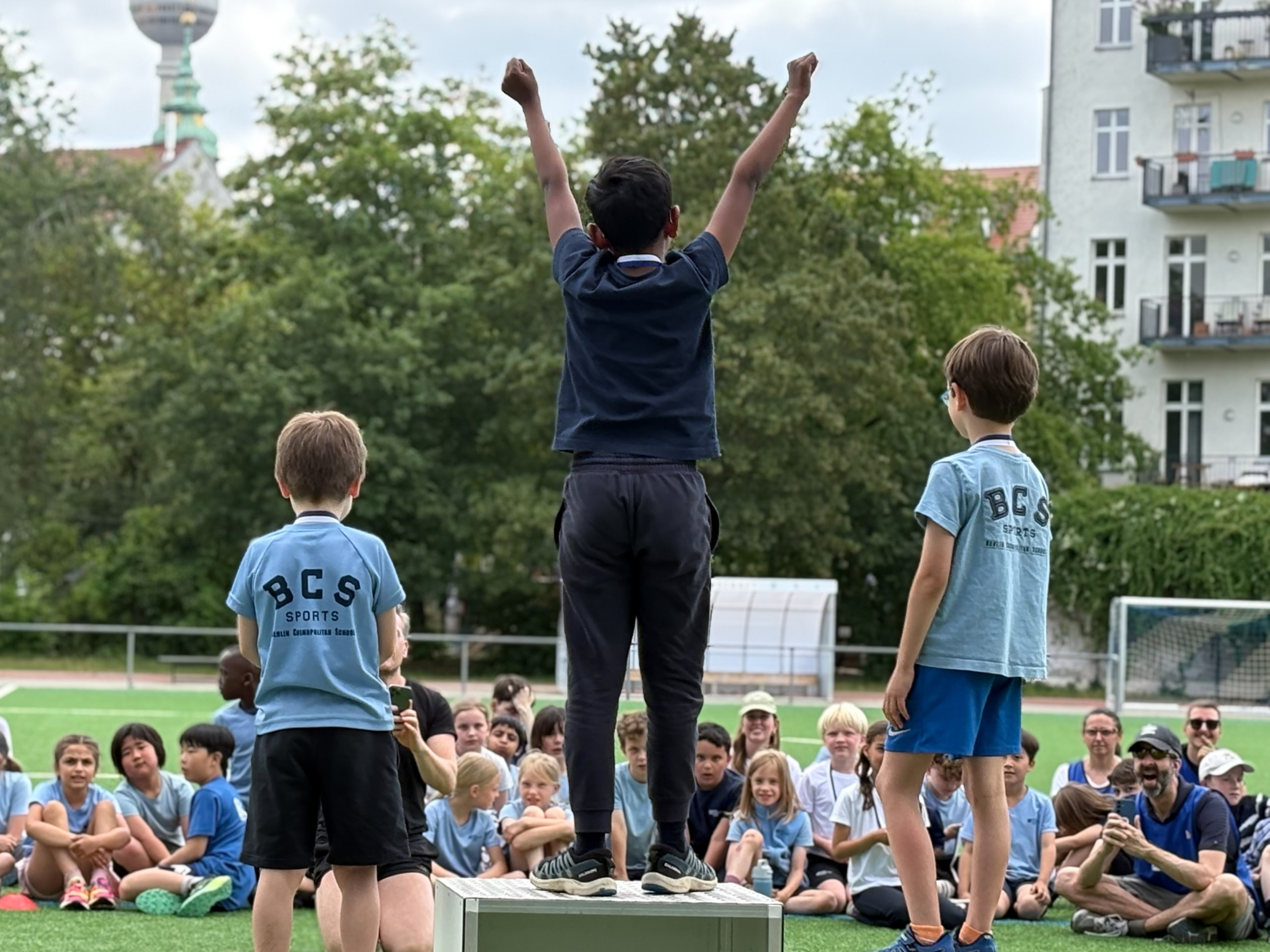 Drei Jungen stehen auf einer Wiese vor einer sitzenden Kindergruppe, der mittlere Junge hebt die Arme nach oben loading=