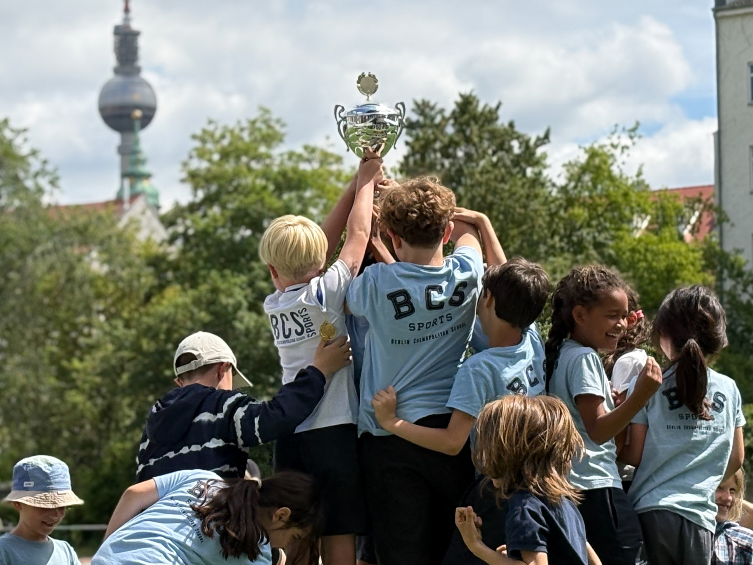 Kinder in Sportkleidung halten gemeinsam einen Pokal hoch vor einer Stadtkulisse mit Fernsehturm loading=