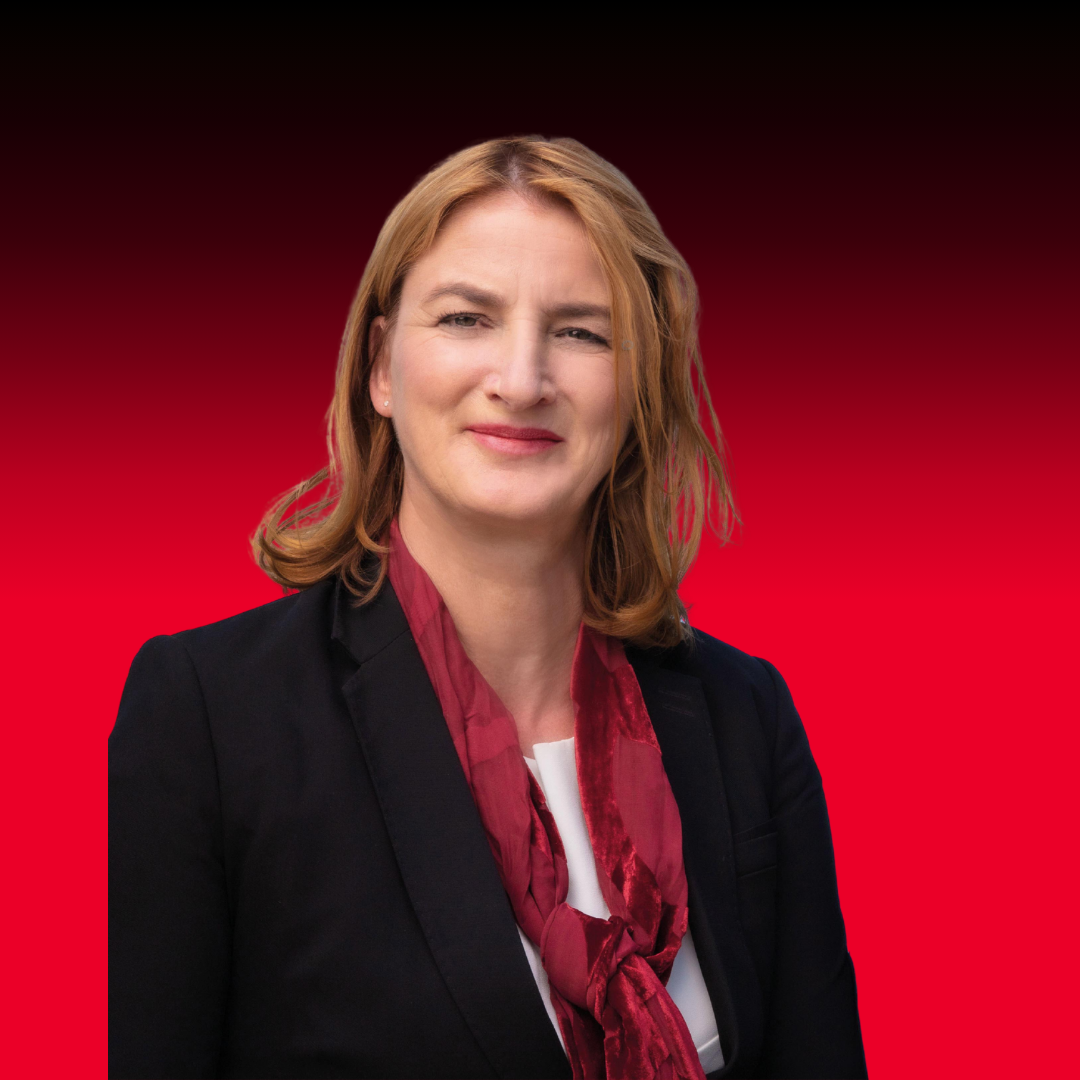 Frau mit schulterlangem, blondem Haar trägt schwarzen Blazer, weiße Bluse und roten Schal vor rotem Hintergrund loading=