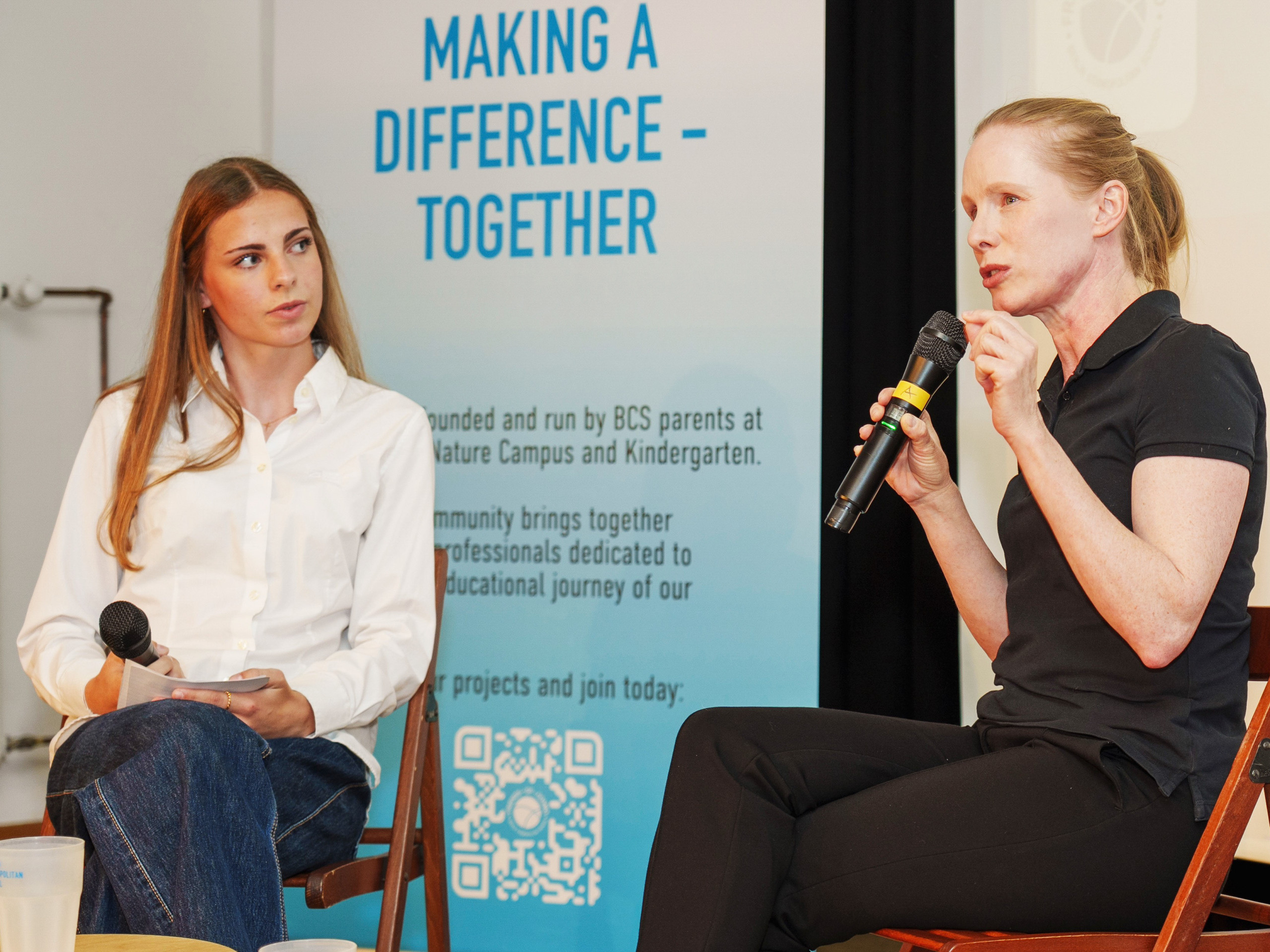 Zwei Frauen sitzen auf Stühlen, eine spricht in ein Mikrofon, die andere hält ein Mikrofon und schaut sie an, im Hintergrund ein Banner mit dem Text 'MAKING A DIFFERENCE - TOGETHER'