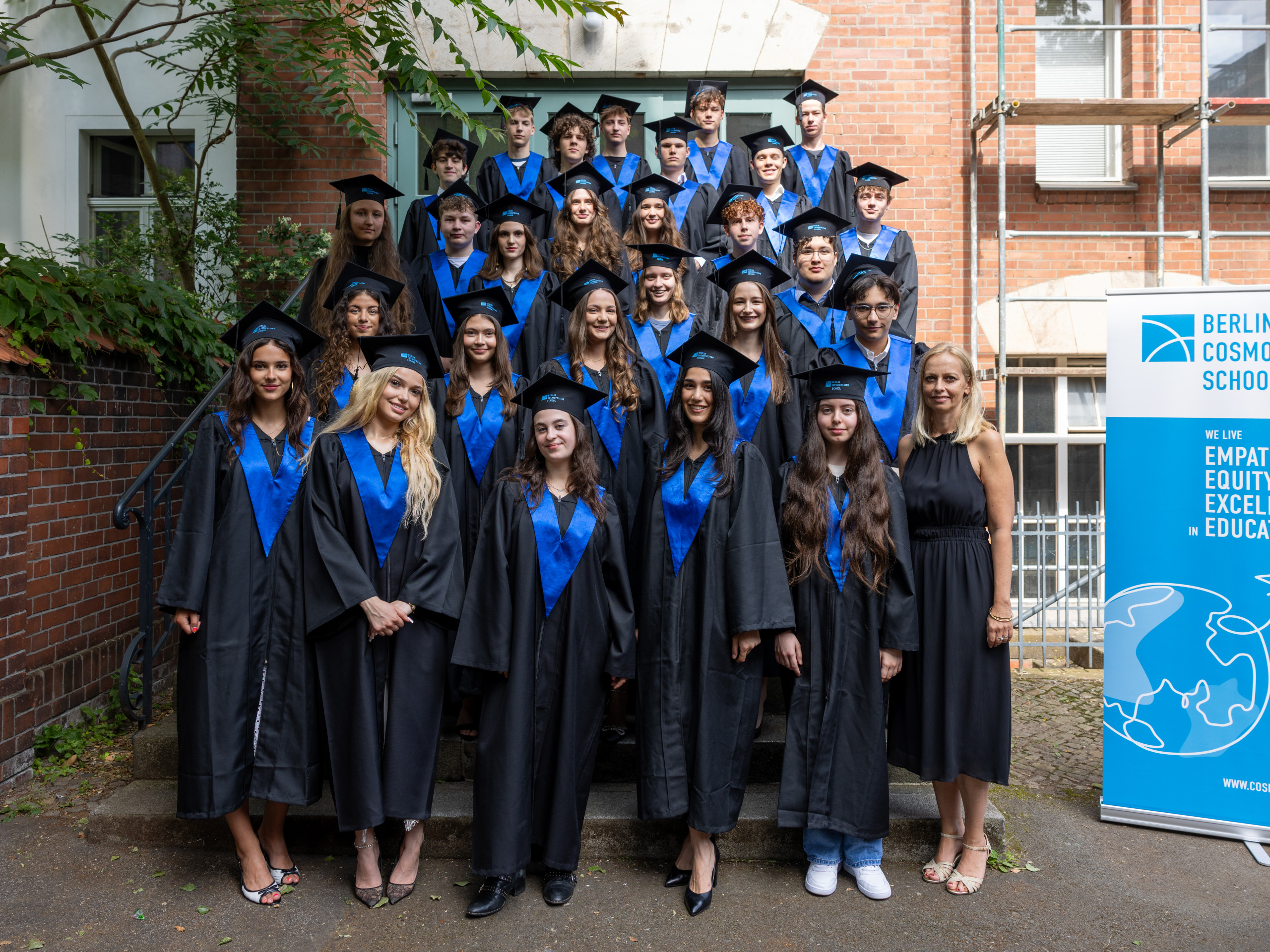 Gruppe von jungen Erwachsenen in schwarzen Talaren mit blauen Schärpen und schwarzen Baretten steht vor einem Backsteingebäude neben einem Banner der Berlin Cosmopolitan School