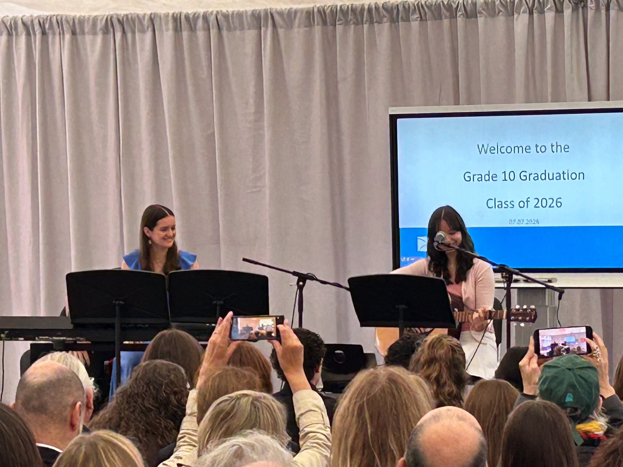 Zwei junge Frauen sitzen auf einer Bühne hinter Notenständern, eine spielt Gitarre, Publikum filmt mit Smartphones, im Hintergrund Leinwand mit Text zur Abschlussfeier der 10. Klasse. loading=