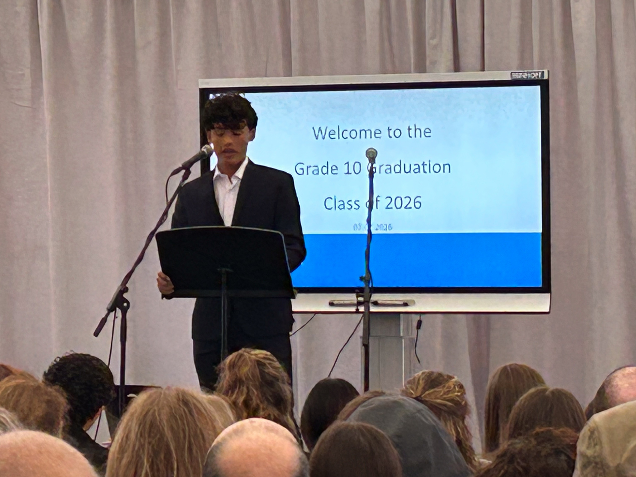 Ein junger Mann in dunklem Anzug steht an einem Rednerpult vor einem Publikum und spricht, im Hintergrund ein Bildschirm mit der Textanzeige 'Welcome to the Grade 10 Graduation Class of 2026' loading=