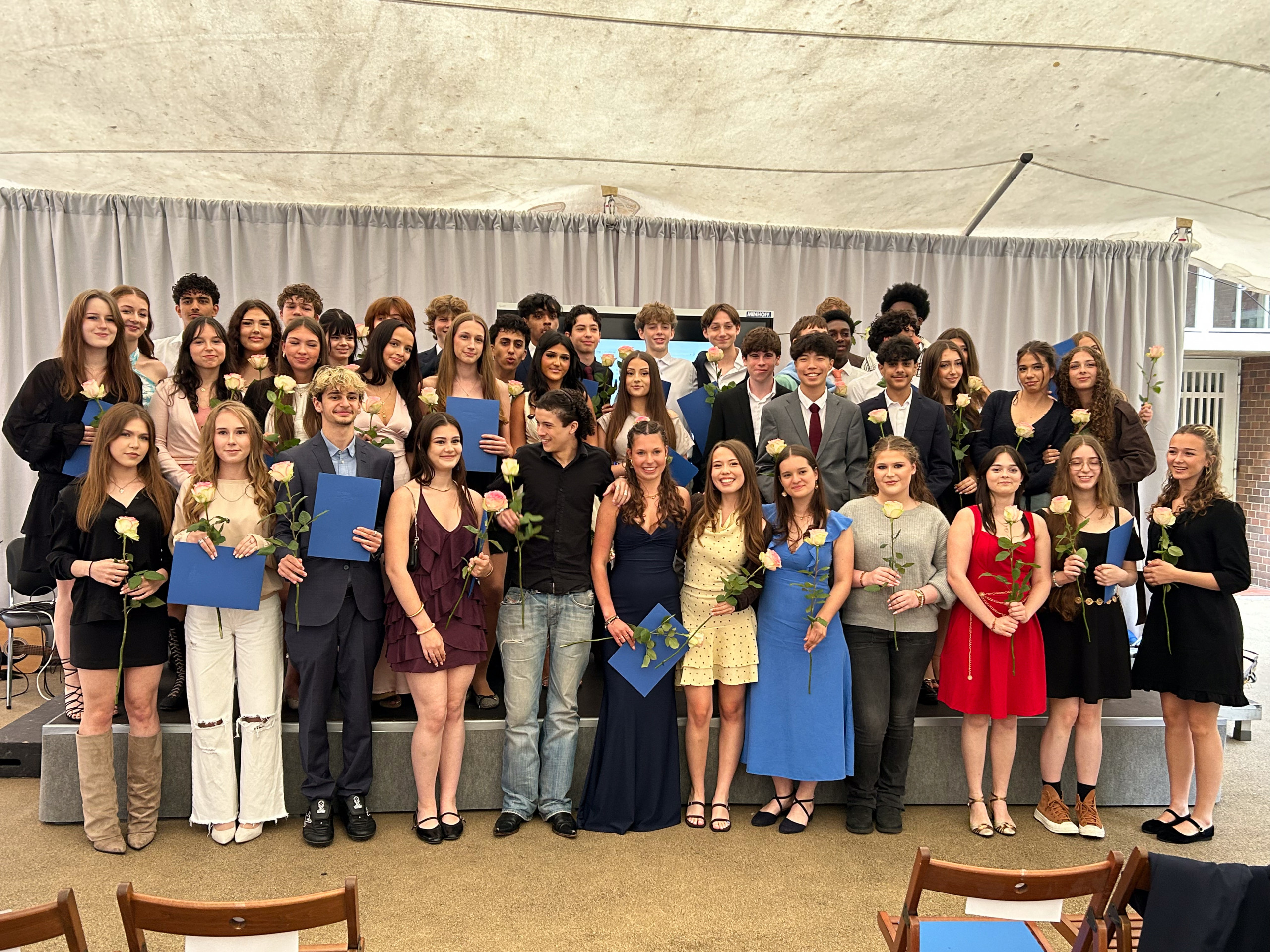 Gruppenfoto von jungen Erwachsenen in festlicher Kleidung, viele halten blaue Ordner und einzelne weiße oder rote Rosen, im Hintergrund ein heller Vorhang unter einem Zelt loading=