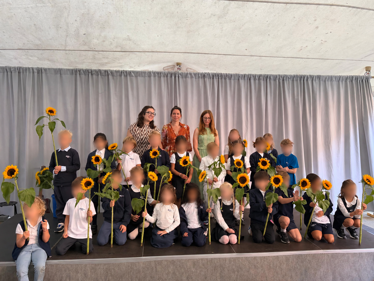 Gruppe von Kindern mit Sonnenblumen auf einer Bühne vor grauem Vorhang, drei Erwachsene stehen hinten loading=