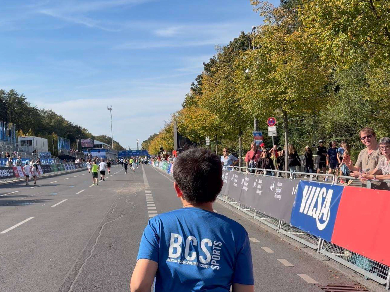 Person von hinten mit blauem T-Shirt mit Aufdruck "BCS SPORTS" auf einer breiten Straße mit weiteren Läufern und Zuschauern an der Seite bei sonnigem Wetter loading=