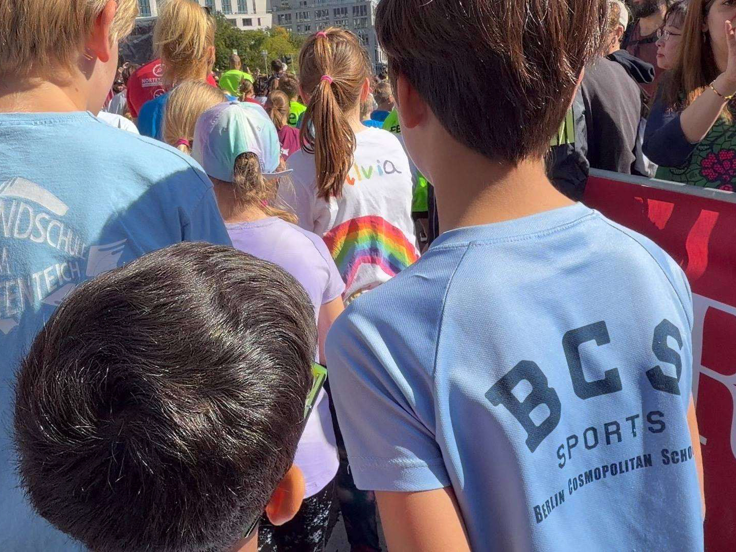 Rückansicht mehrerer Kinder in einer Menschenmenge, zwei tragen hellblaue T-Shirts mit der Aufschrift 'BCS SPORTS BERLIN COSMOPOLITAN SCH'.