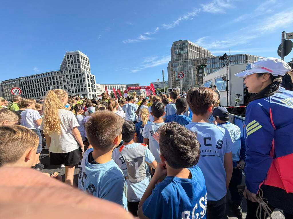 Gruppe von Kindern und Jugendlichen steht auf einer Straße vor modernen Gebäuden, eine Person in blauer Uniform mit weißem Helm steht rechts